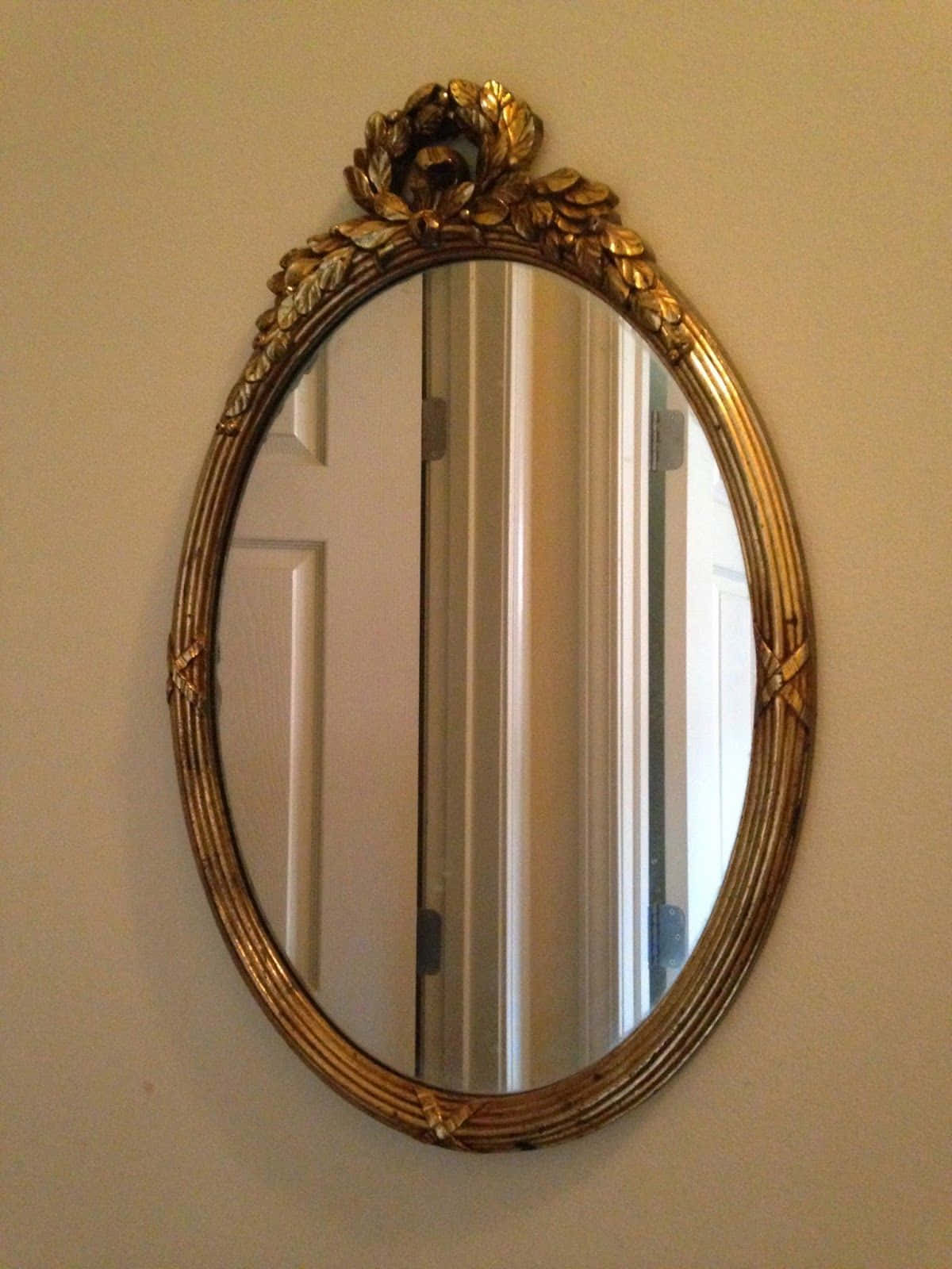 mirror 桌布 壁紙 — 一個帶有葉狀頂飾的華麗金色橢圓鏡子掛在米色牆上，反射出白色門和走廊。