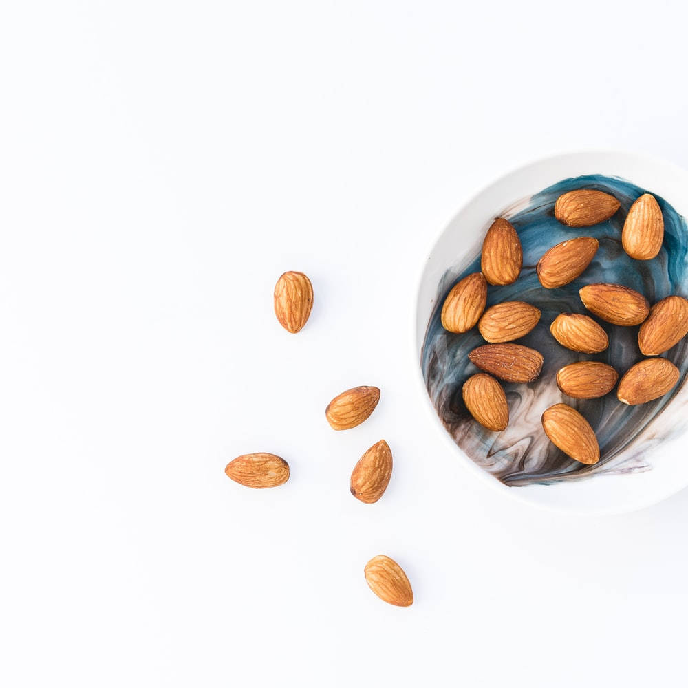 Almond 桌布 壁紙 — 一個帶有藍色和灰色漩渦的白色碗裡裝著杏仁，有幾顆散落在乾淨的白色表面上。