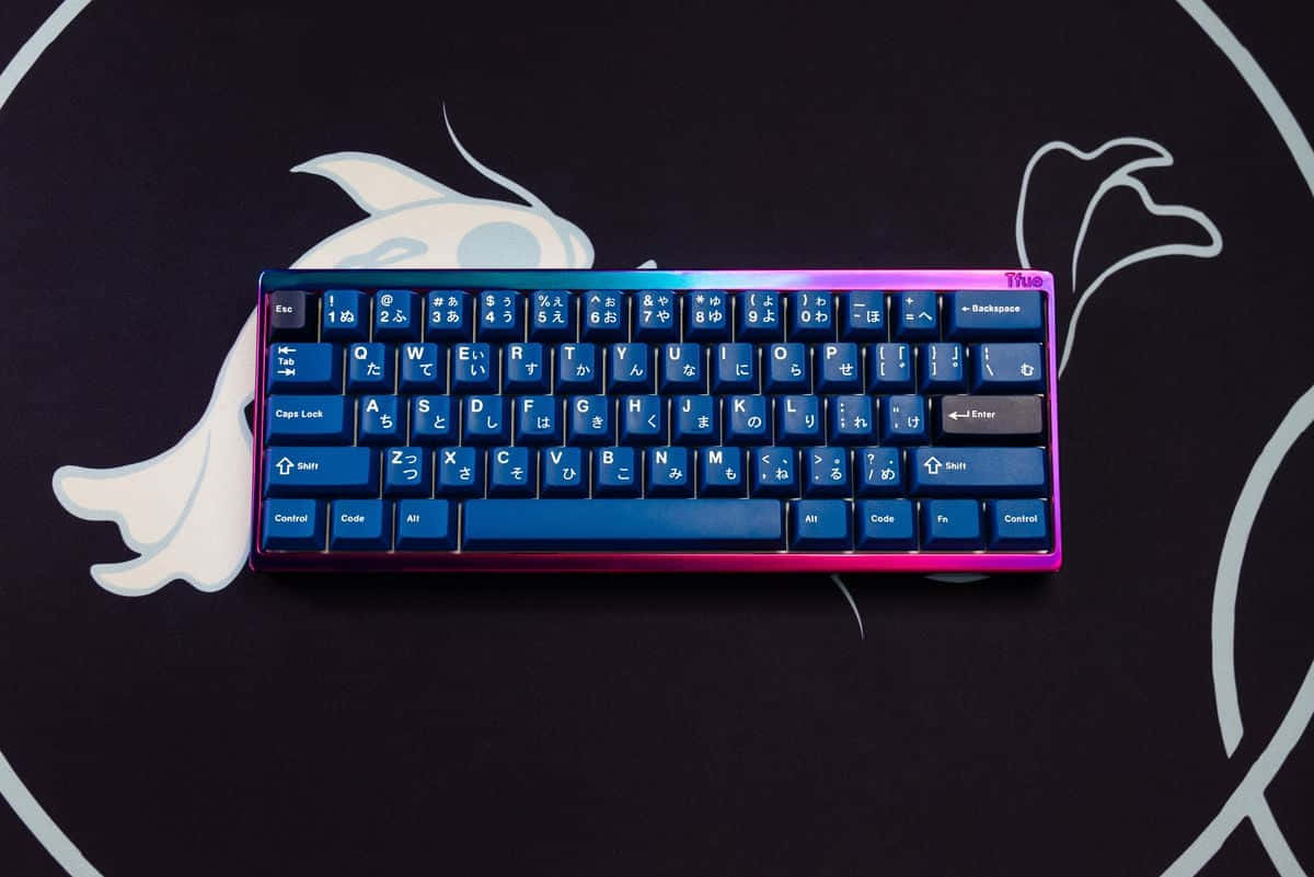 Cute Keyboard 桌布 壁紙 — 一個帶有藍色和粉紅色漸變按鍵的緊湊型鍵盤，放置在帶有白色幽靈生物輪廓的黑色表面上。