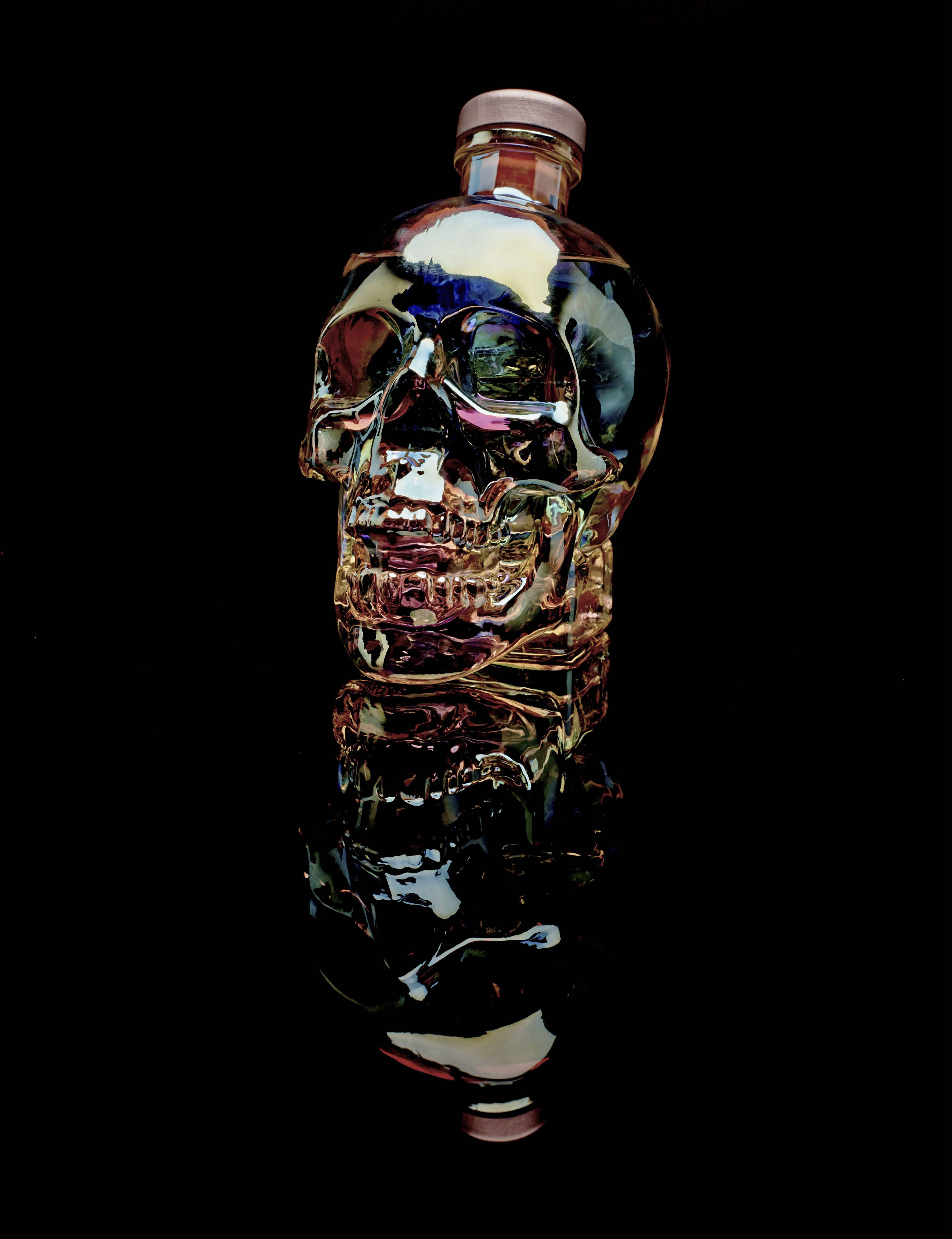 Drink 桌布 壁紙 — 一個帶有虹彩金屬反光的光澤骷髏形瓶子，矗立在黑色背景和下方的鏡面之上。
