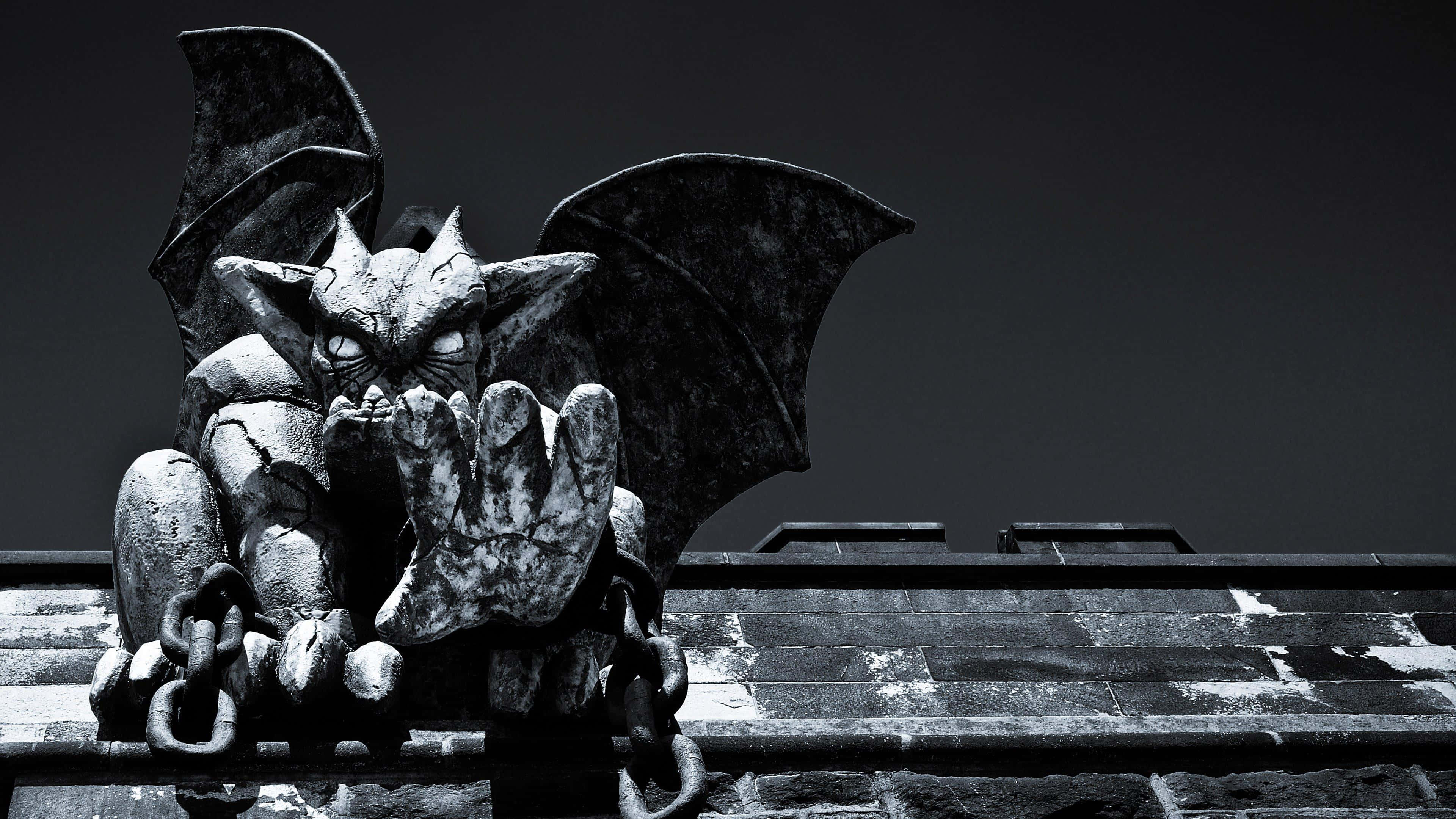 Gargoyle 桌布 壁紙 — 一個帶有蝙蝠翅膀和利爪的可怕石像鬼雕像，棲息在石屋頂上，背景是黑暗的天空。