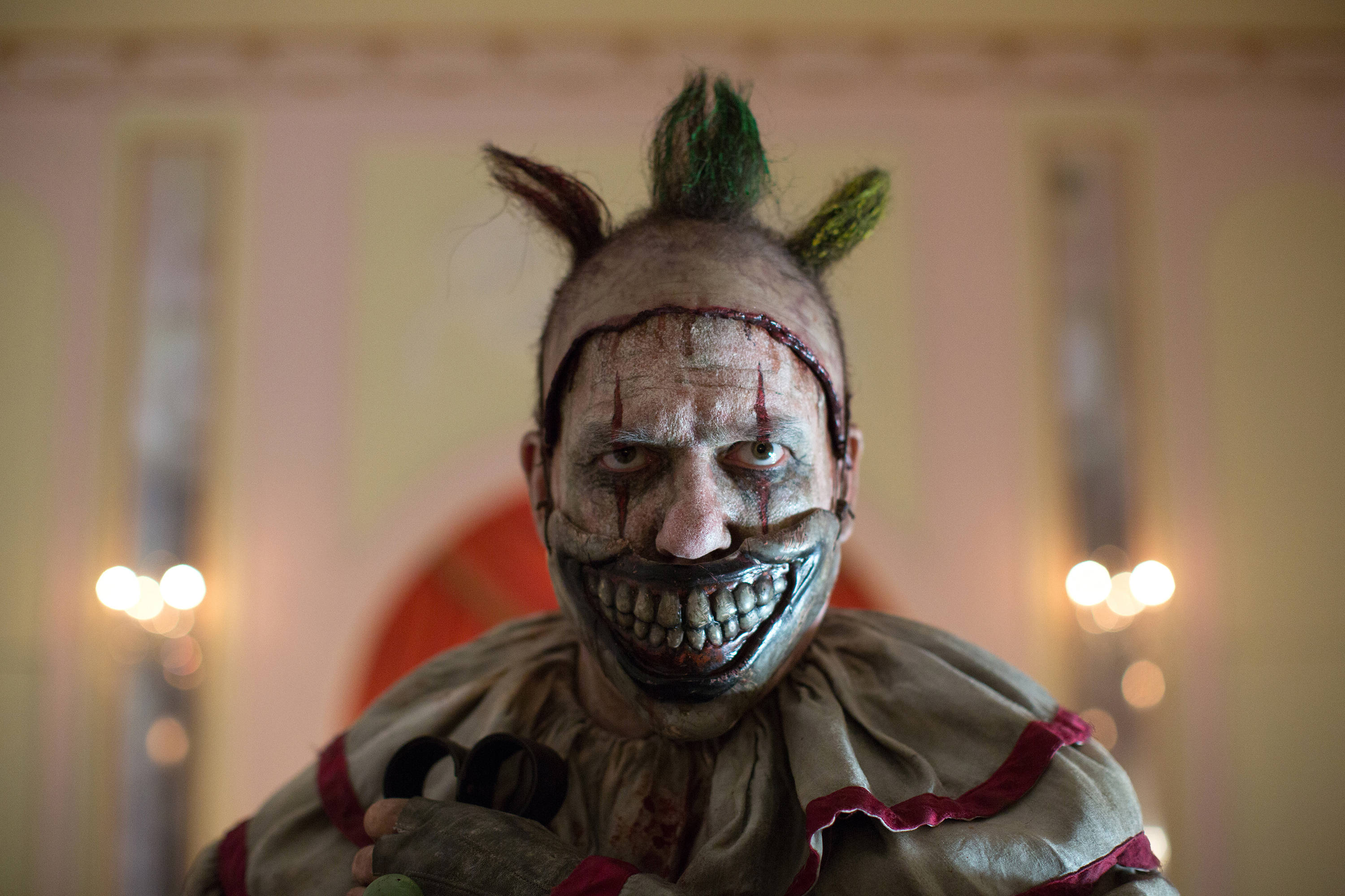 American Horror Story 桌布 壁紙 — 一個帶有血跡化妝和綠色莫西干頭的威脅性小丑，在昏暗的房間裡凝視著相機。