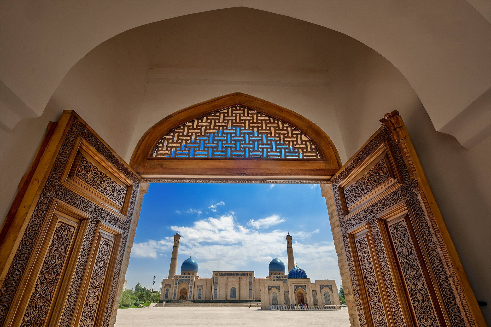 Uzbekistan 桌布 壁紙 — 一個帶有複雜木雕的大型拱形門口，框住了一座在明亮藍天下擁有藍色圓頂的雄偉清真寺。