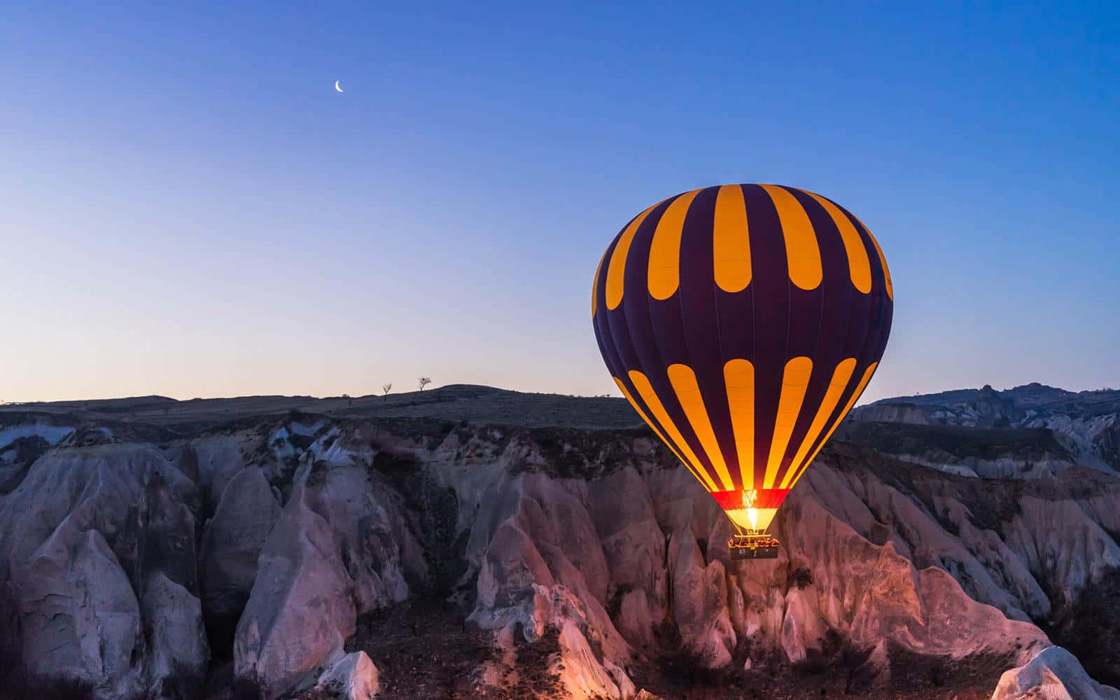 Hot Air Balloon 桌布 壁紙 — 一個帶有醒目黃色和紫色條紋的熱氣球，在暮色天空中，新月懸掛在超現實的岩石 formations 之上，溫暖地發光。