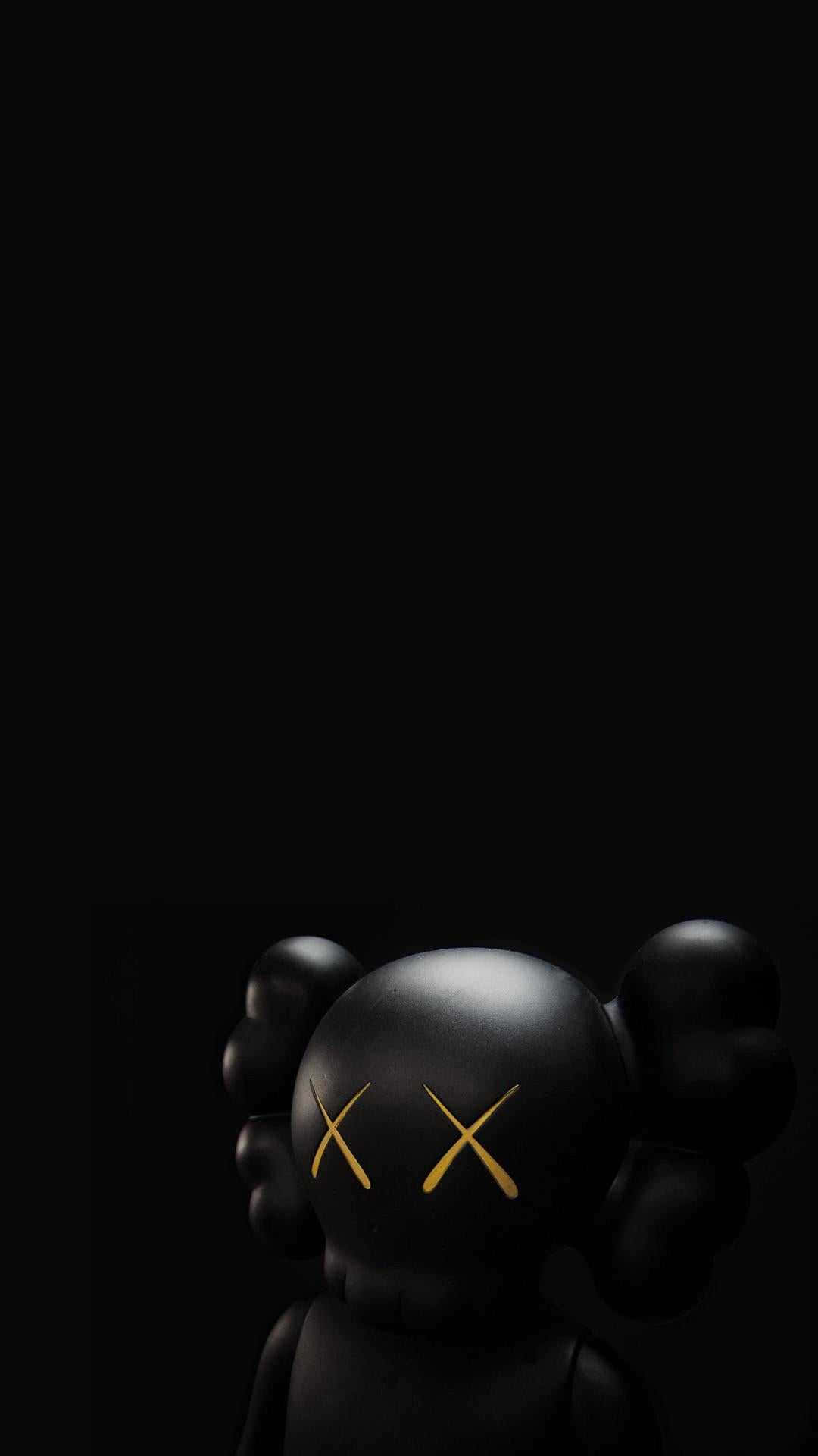 kaws 4K 桌布 壁紙 — 一個帶有金色X眼睛的光澤黑色KAWS人偶，在純黑色背景的深陰影中站立。