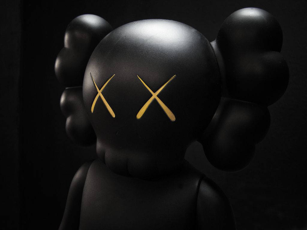 KAWS 桌布 壁紙 — 一個帶有金色X眼睛的光澤黑色KAWS人偶站在陰影黑暗中，其標誌性的氣球狀耳朵被柔和照亮。