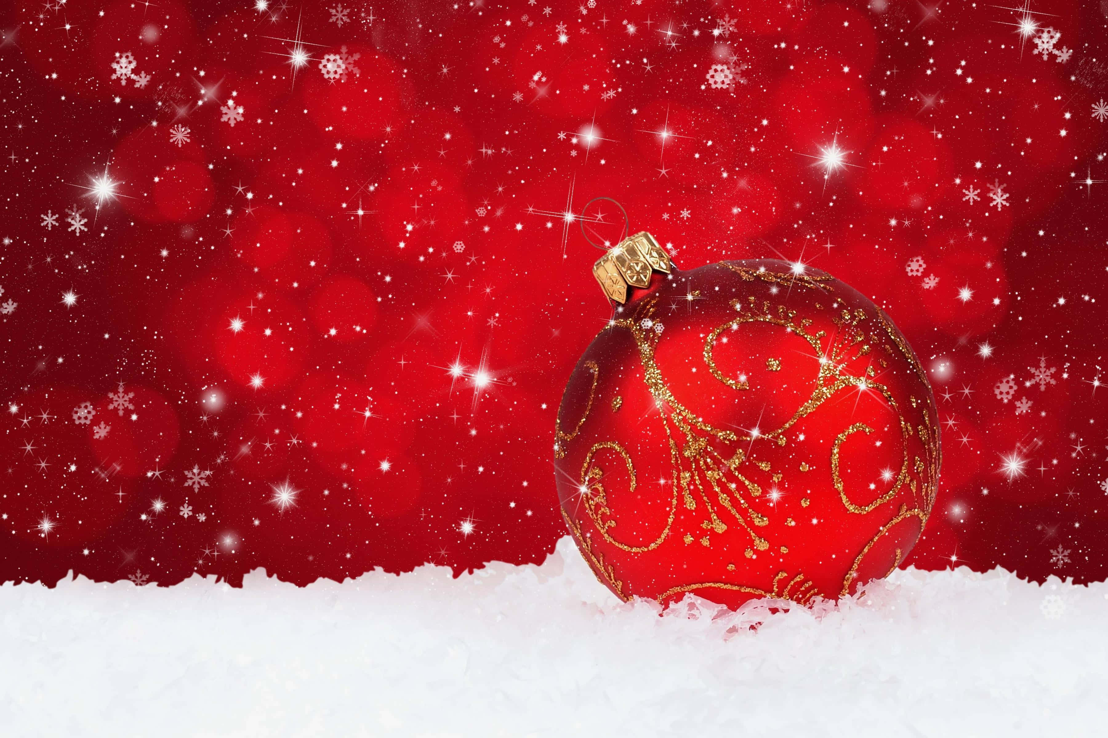 Snowy Christmas 桌布 壁紙 — 一個帶有金色漩渦的光滑紅色聖誕裝飾品，躺在蓬鬆的雪中，背景是充滿活力的紅色散景和閃爍的雪花。