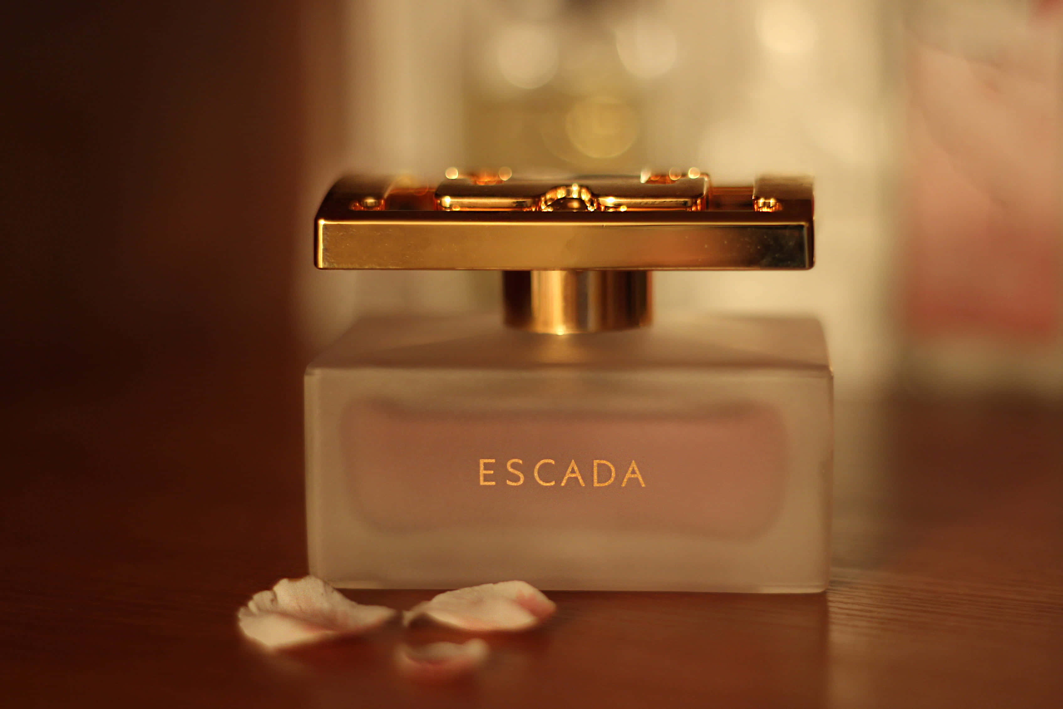 粉紅色系 桌布 壁紙 — 一個帶有金色瓶蓋的奢華 Escada 香水瓶放置在木質表面上，周圍環繞著精緻的玫瑰花瓣。