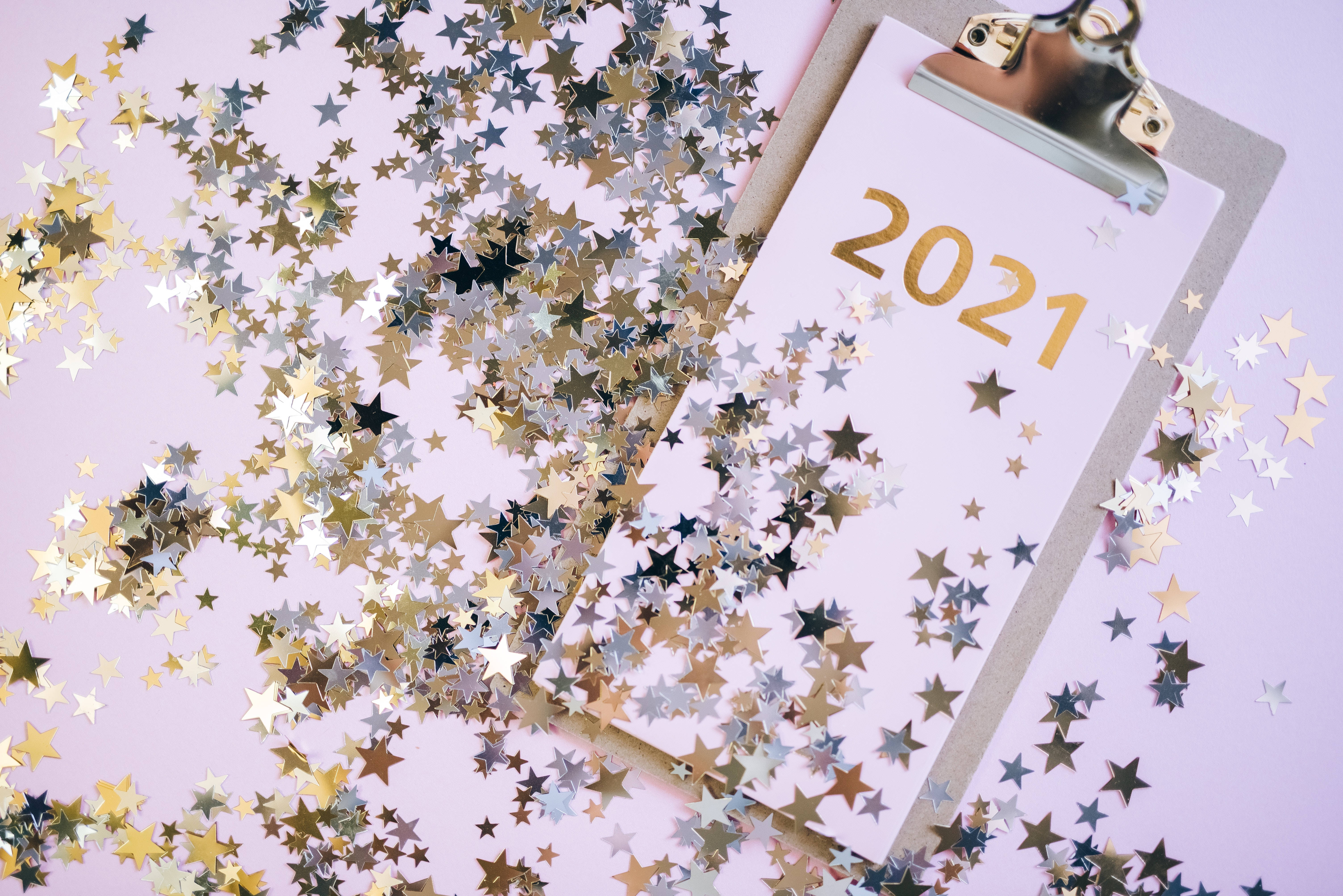 Confetti 桌布 壁紙 — 一個帶有金色箔紙 '2021' 的剪貼板，周圍散落著金色和銀色的星星紙屑，背景為柔和的粉紅色。
