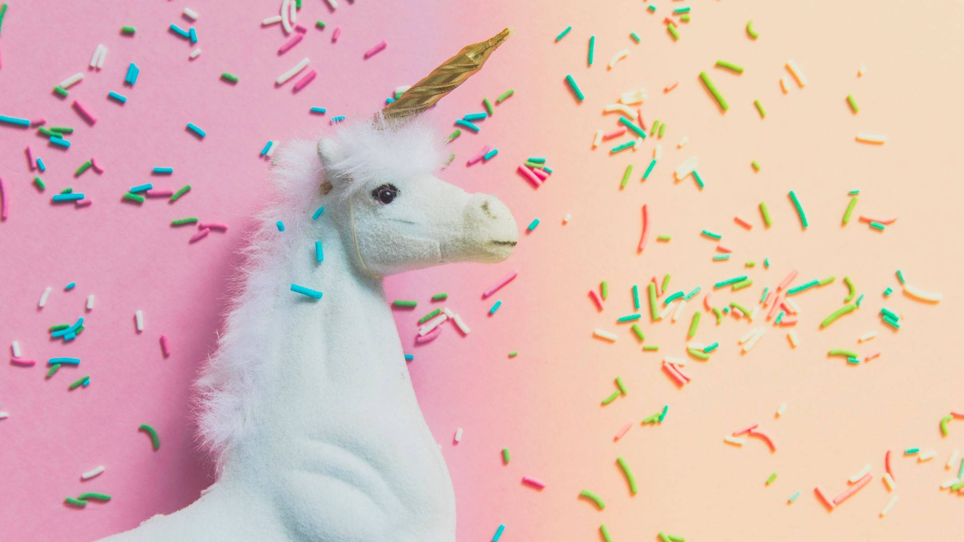 Cool Unicorn 桌布 壁紙 — 一個帶有金色角的毛絨白色獨角獸頭，坐在粉紅到蜜桃漸變背景上的彩色糖針之間。