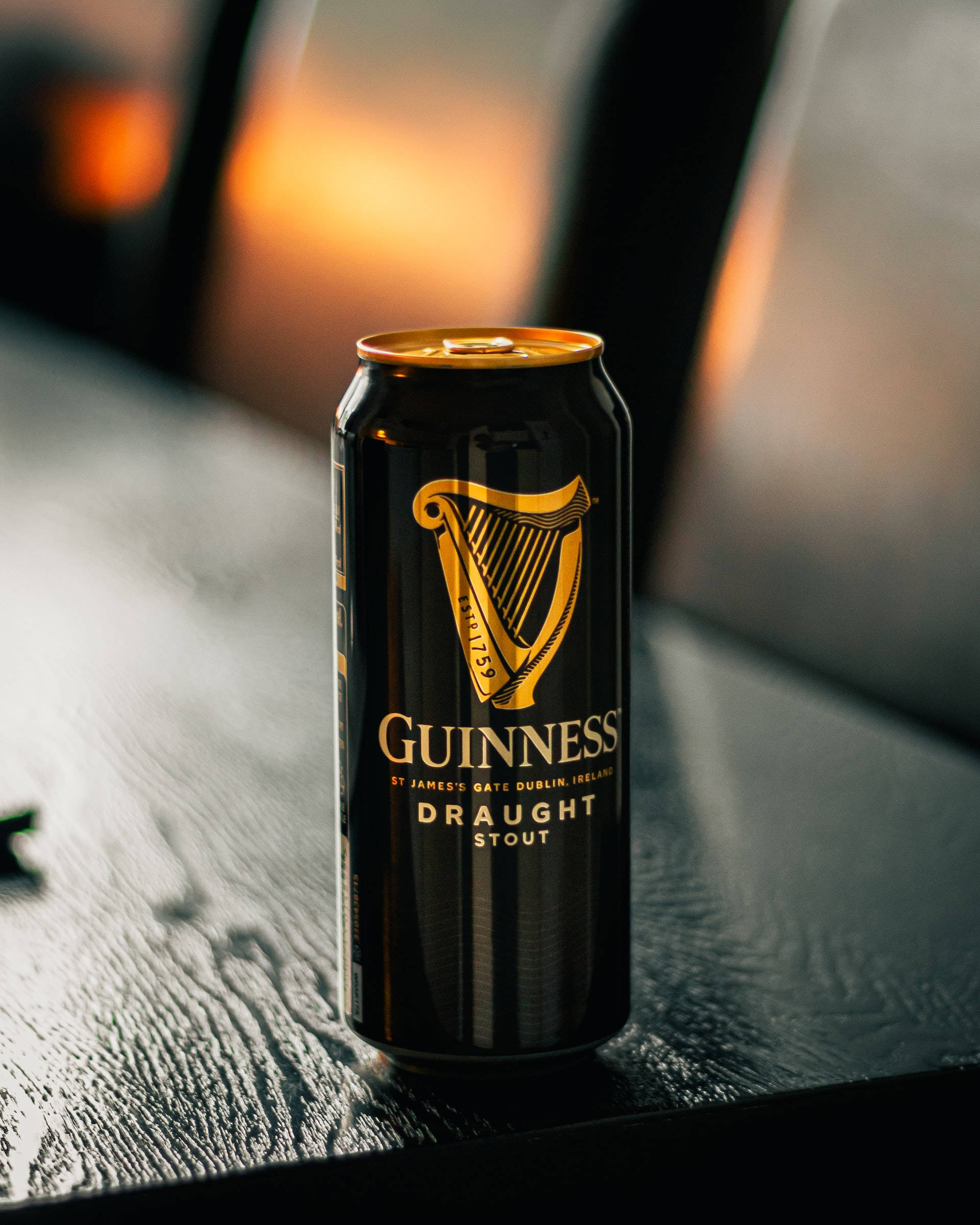 Beer 桌布 壁紙 — 一個帶有金色豎琴標誌的黑色健力士生啤黑啤罐，在溫暖的環境燈光下放在一張深色紋理桌子上。