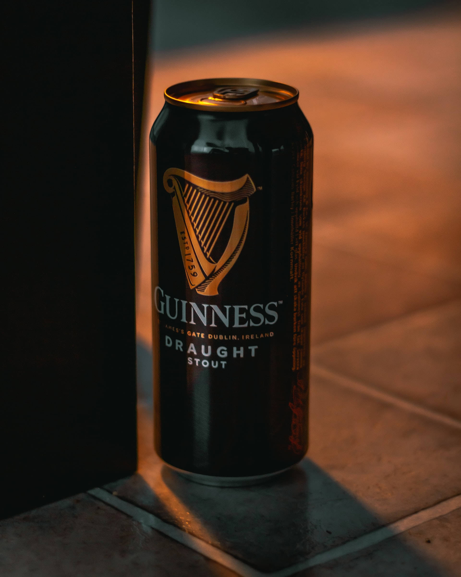 Beer 桌布 壁紙 — 一個帶有金色豎琴標誌的黑色健力士黑啤酒罐，放在沐浴在溫暖金色光線中的瓷磚地板上。