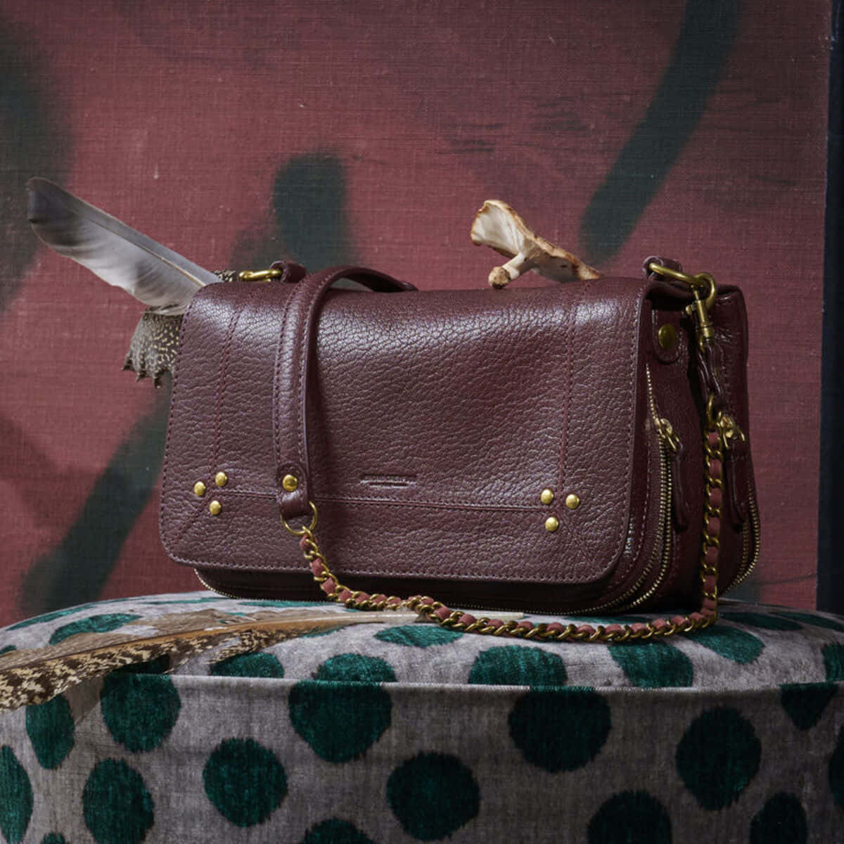 Handbag 桌布 壁紙 — 一個帶有金色金屬配件和羽毛裝飾的勃艮第皮革斜挎包，放在綠色圓點布料上，背景是深紅色紋理牆壁。