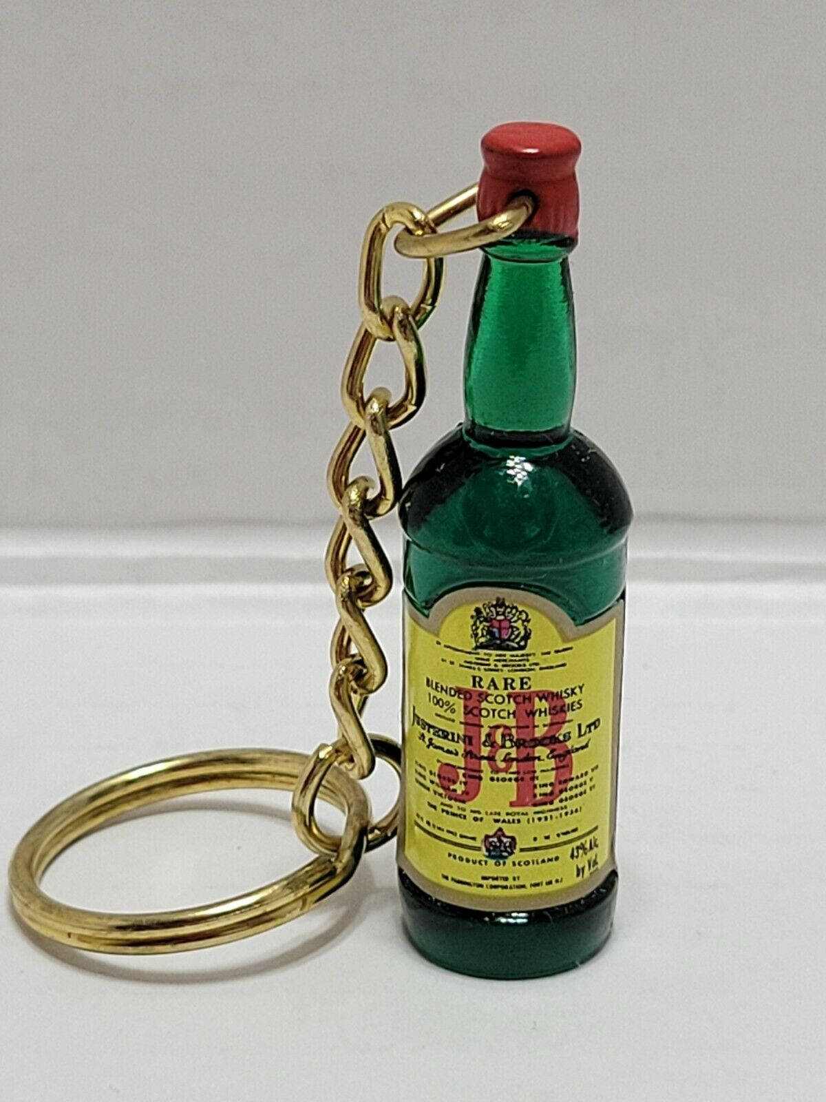drinks 桌布 壁紙 — 一個帶有金色鏈條和紅色蓋子的迷你綠色威士忌酒瓶鑰匙圈放在純白色表面上。