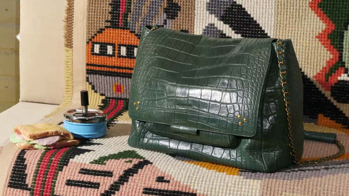 Handbag 桌布 壁紙 — 一個帶有金色鏈條的光澤綠色鱷魚紋手袋放在色彩繽紛的編織布料上，旁邊有一個藍色煙灰缸和三明治。