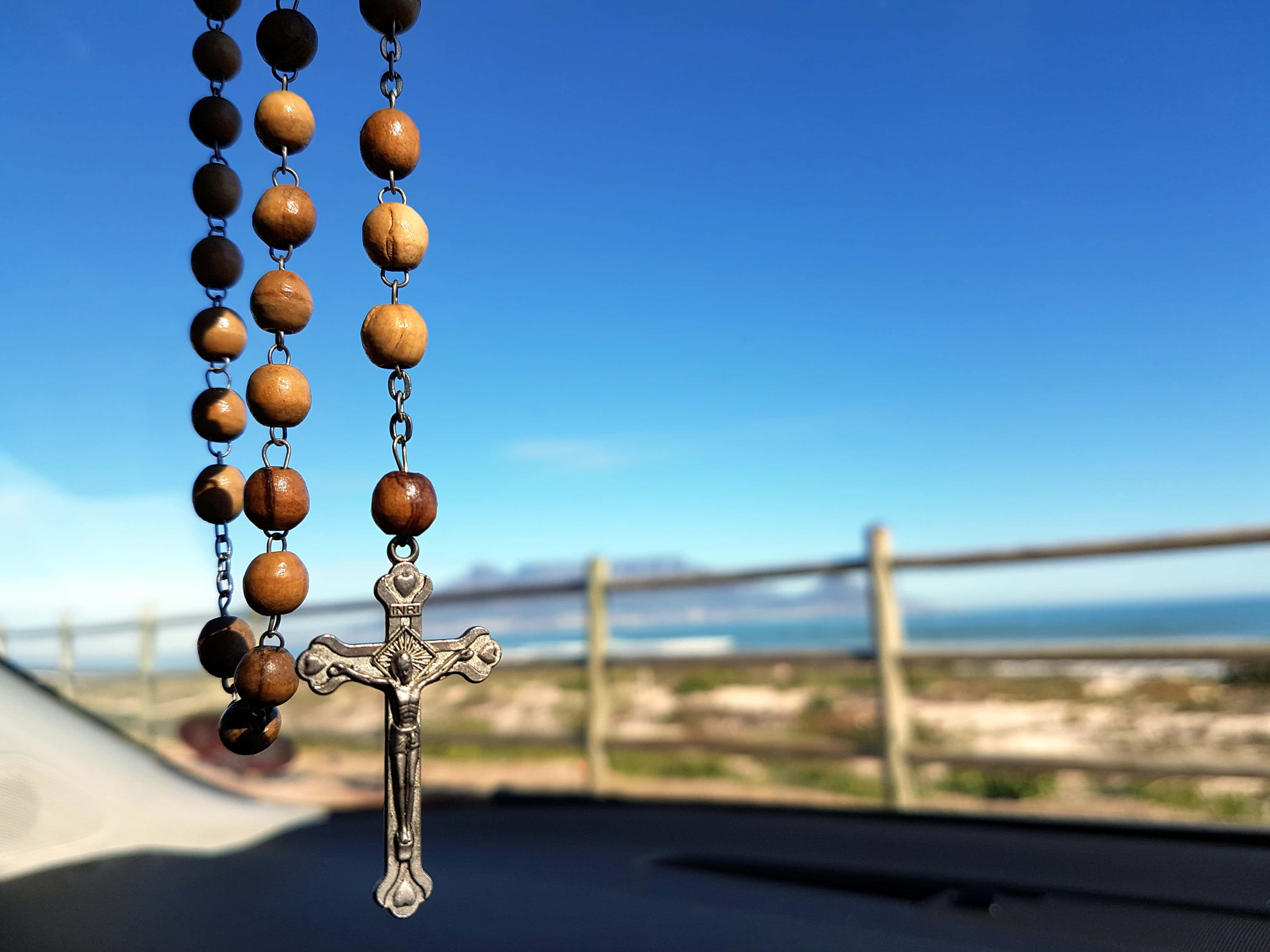 Catholic Desktop 桌布 壁紙 — 一個帶有銀色十字架的木製玫瑰經念珠掛在汽車後視鏡上，背景是晴朗的藍天和海岸景色。