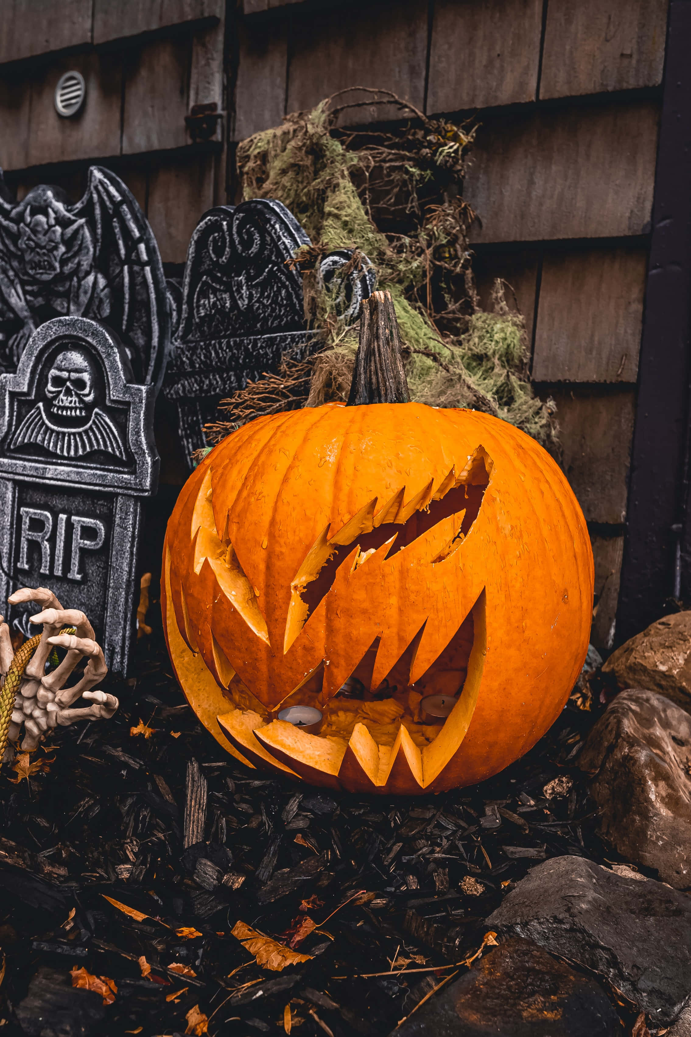 jack o'lantern 桌布 壁紙 — 一個帶有鋸齒狀牙齒的威脅性雕刻南瓜坐在墓碑和長滿苔蘚的木頭之間，營造出詭異的萬聖節場景。