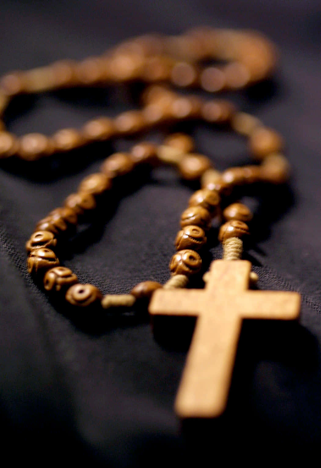 Rosary 桌布 壁紙 — 一個帶有雕刻珠子和十字架的木製玫瑰經念珠，在溫暖燈光下輕輕放在深色布料上。