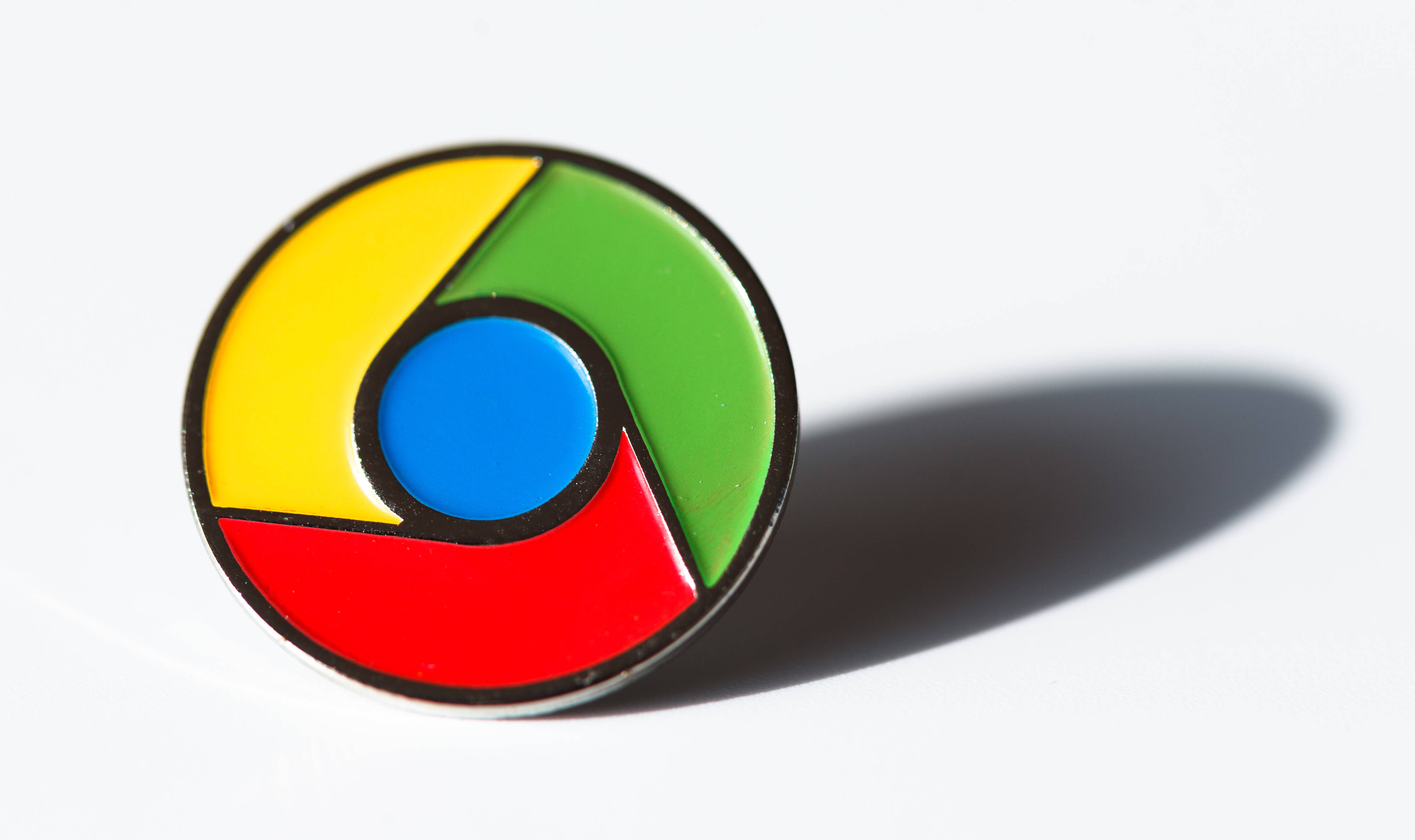 Google Chrome 桌布 壁紙 — 一個帶有黃色、綠色、紅色和藍色部分的光澤圓形 Chrome 標誌別針，在白色表面上投射出柔和陰影。
