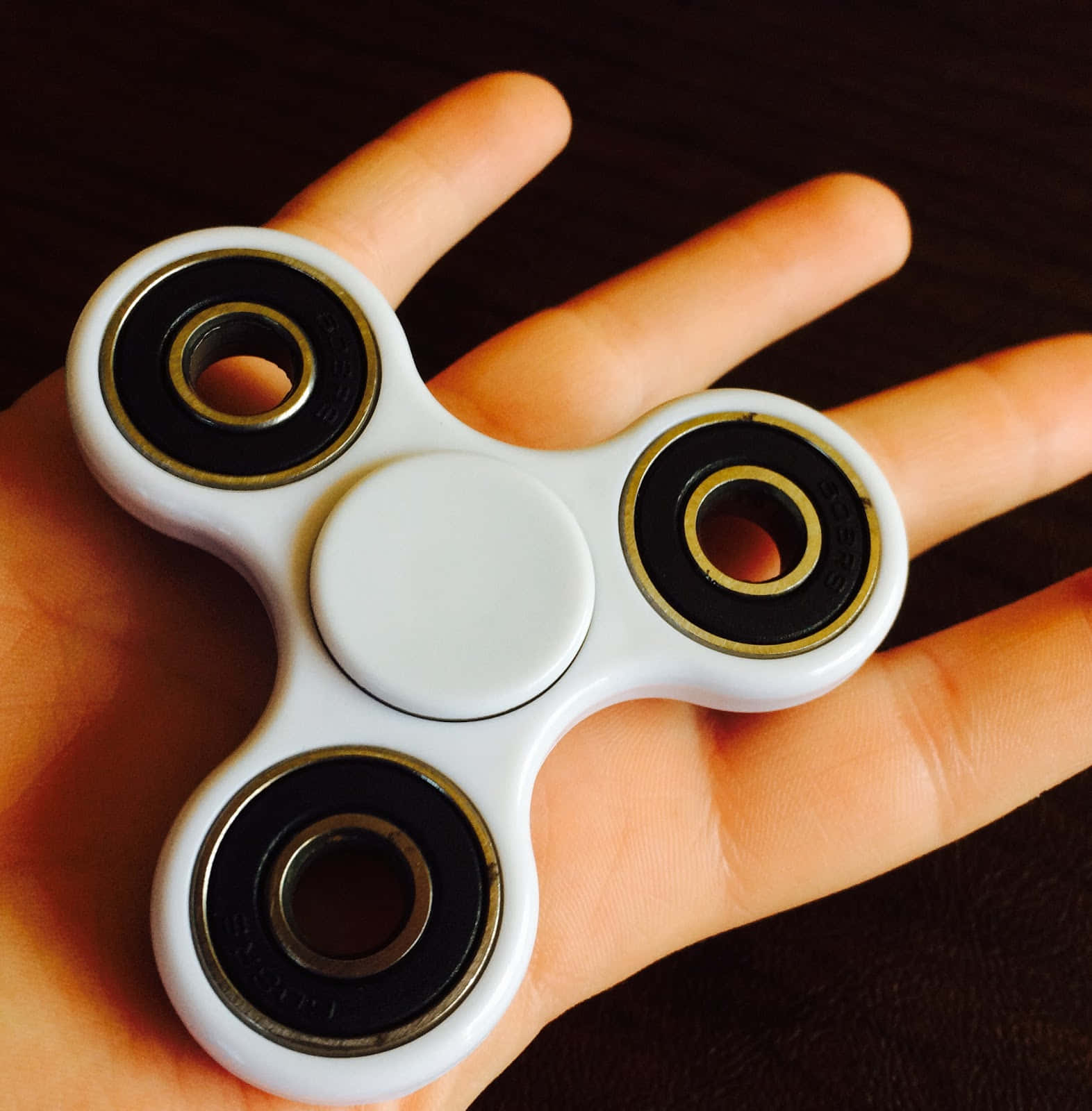 Fidget 桌布 壁紙 — 一個帶有黑色和金色軸承的白色指尖陀螺，放在張開的手掌上，背景是深色木質表面。