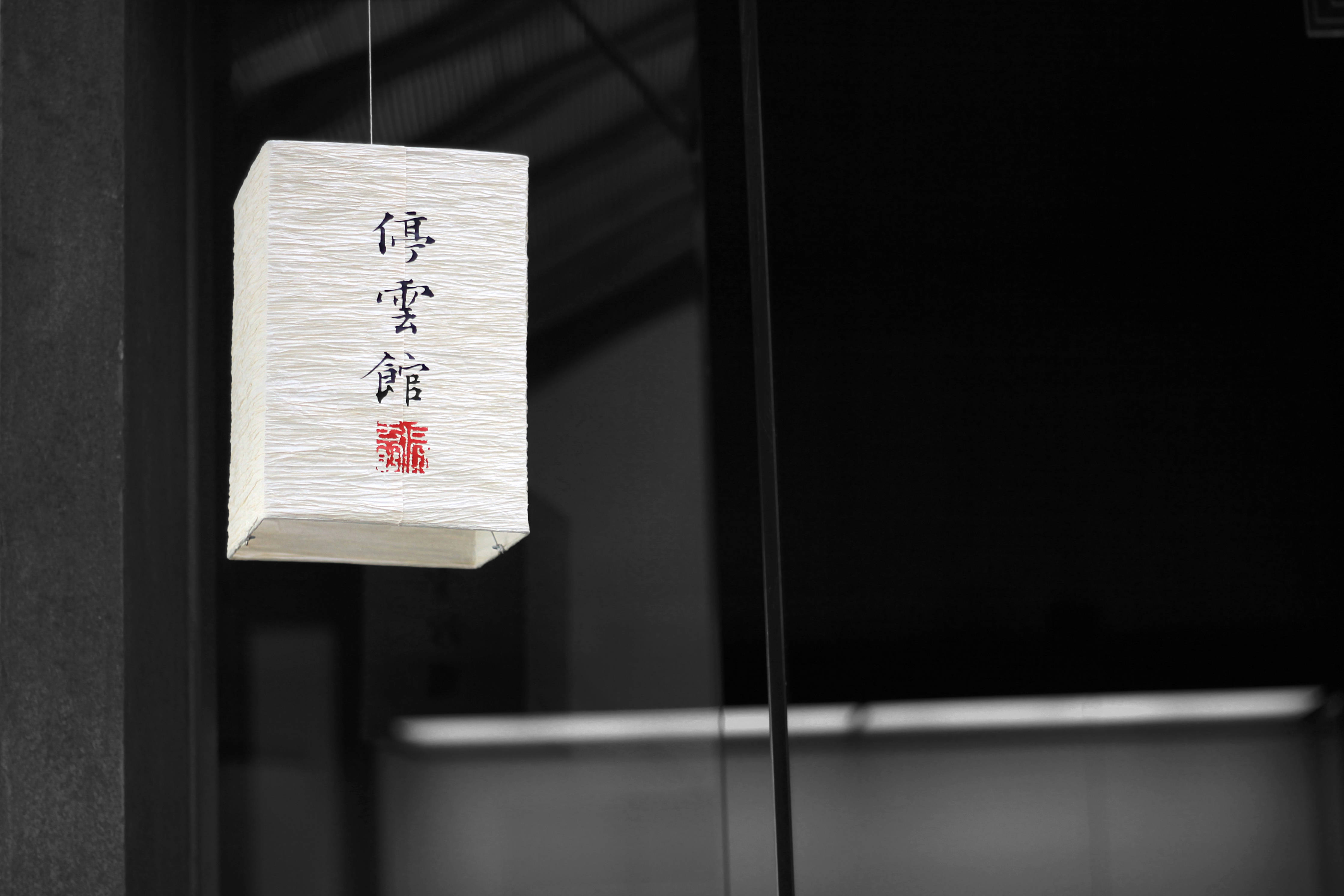 中文 桌布 壁紙 — 一個帶有黑色書法和紅色印章的白色日本紙燈籠懸掛在黑暗的現代室內空間中。