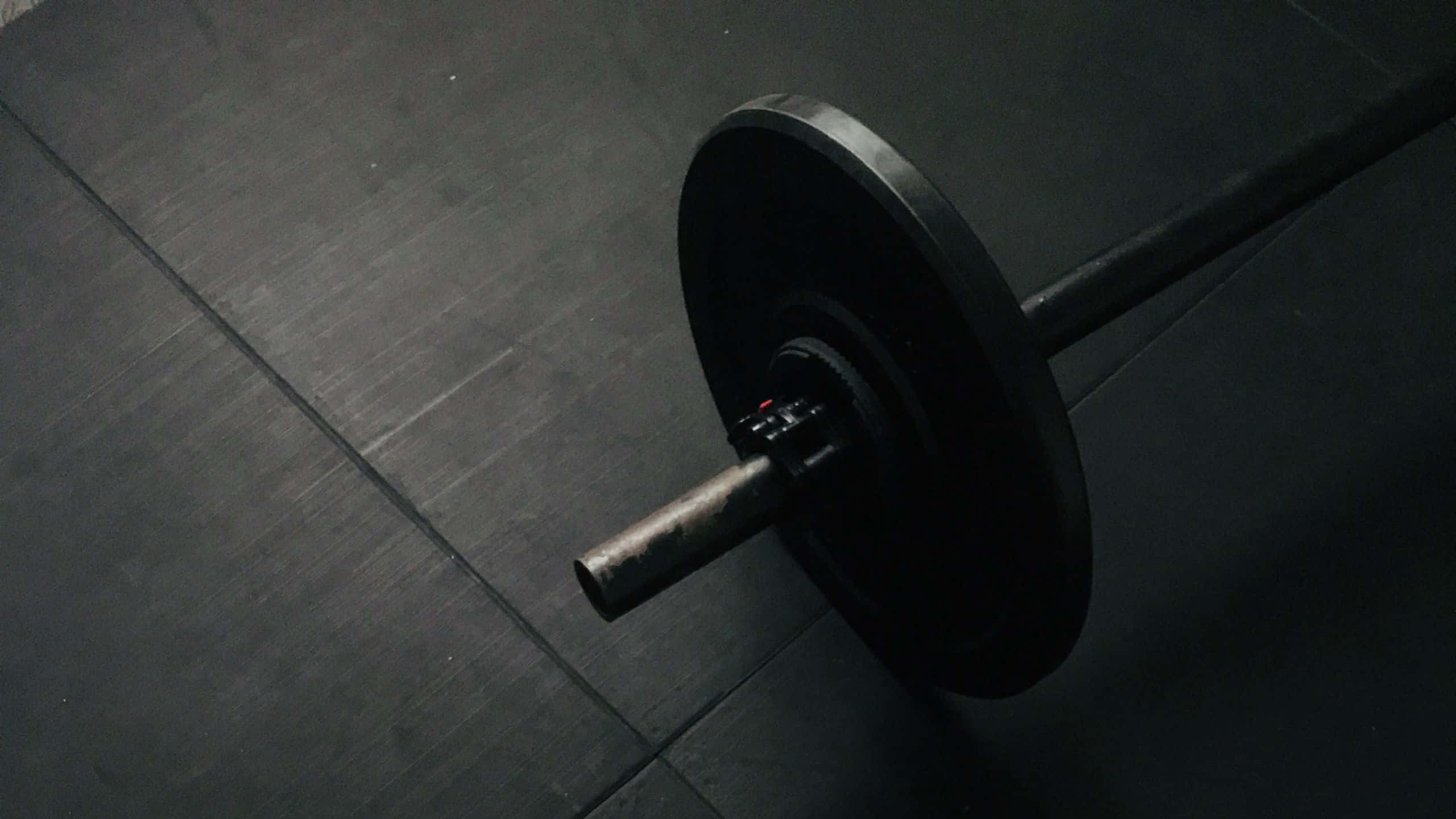 Barbell 桌布 壁紙 — 一個帶有黑色槓鈴片的沉重槓鈴，在戲劇性的低光下放置在深色健身房地板上。