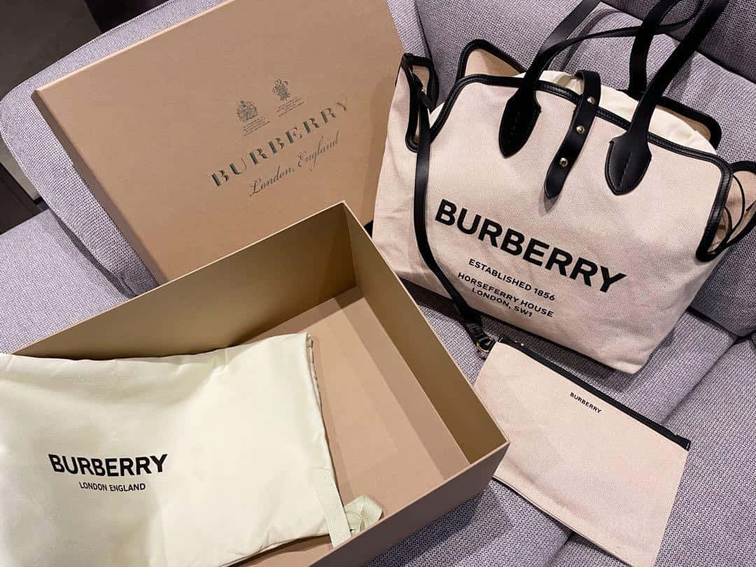 Burberry 桌布 壁紙 — 一個帶有黑色皮革飾邊的 Burberry 帆布手提袋，旁邊放著其品牌盒子和防塵袋，位於灰色紋理沙發上。