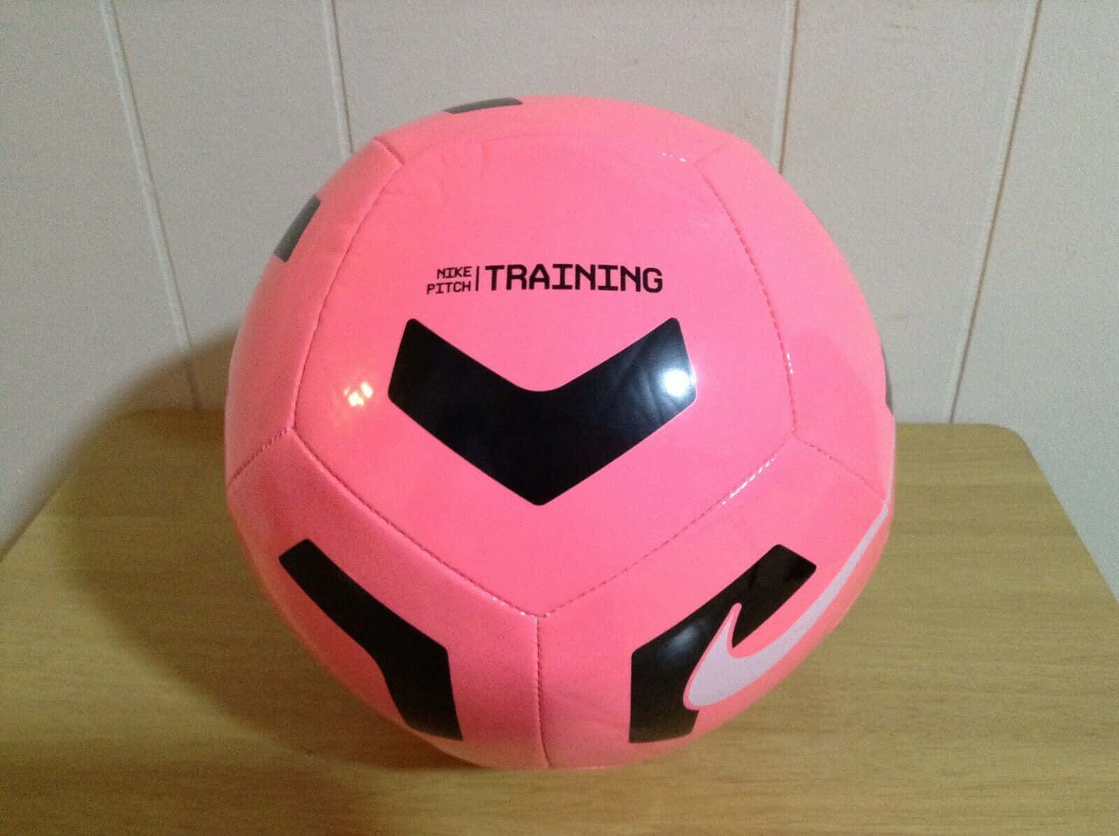 Pink Soccer Ball 桌布 壁紙 — 一個帶有黑色點綴的亮粉紅色Nike訓練足球放在白色鑲板牆前的木質表面上。