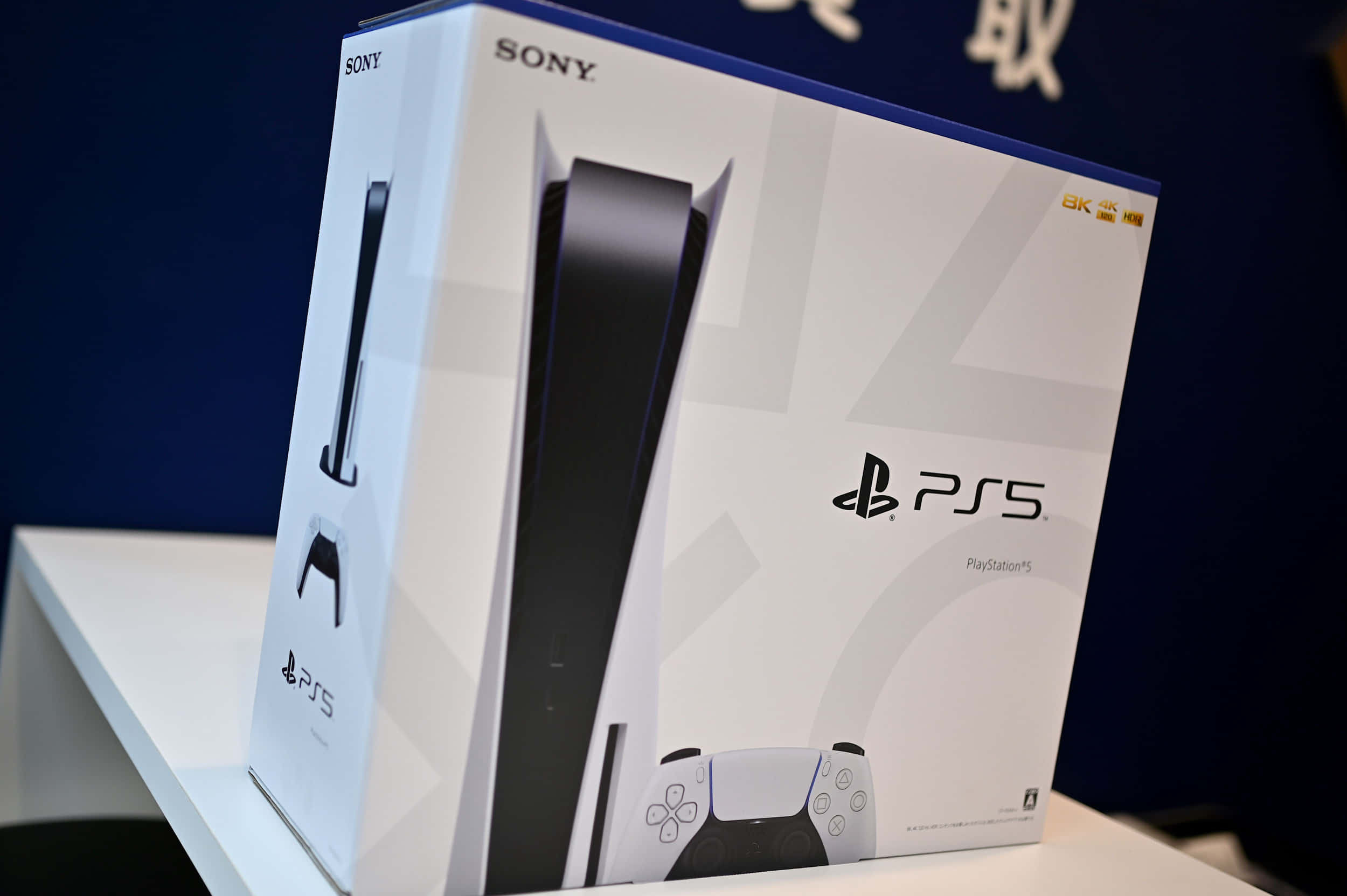 PlayStation 桌布 壁紙 — 一個帶有黑色點綴的白色PlayStation 5主機盒放在白色展示架上，背景為深藍色。
