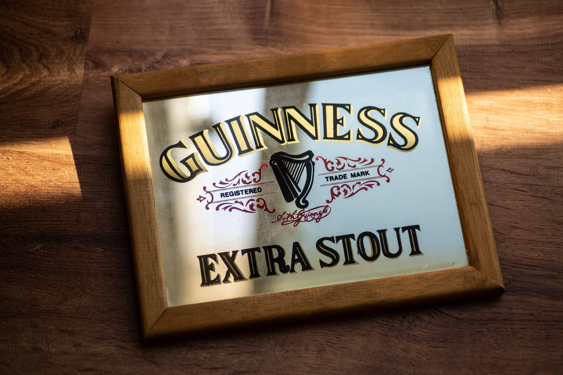Drink 桌布 壁紙 — 一個帶框的健力士Extra Stout鏡面標誌在陽光照射的木桌上閃耀著金色字體和豎琴標誌。