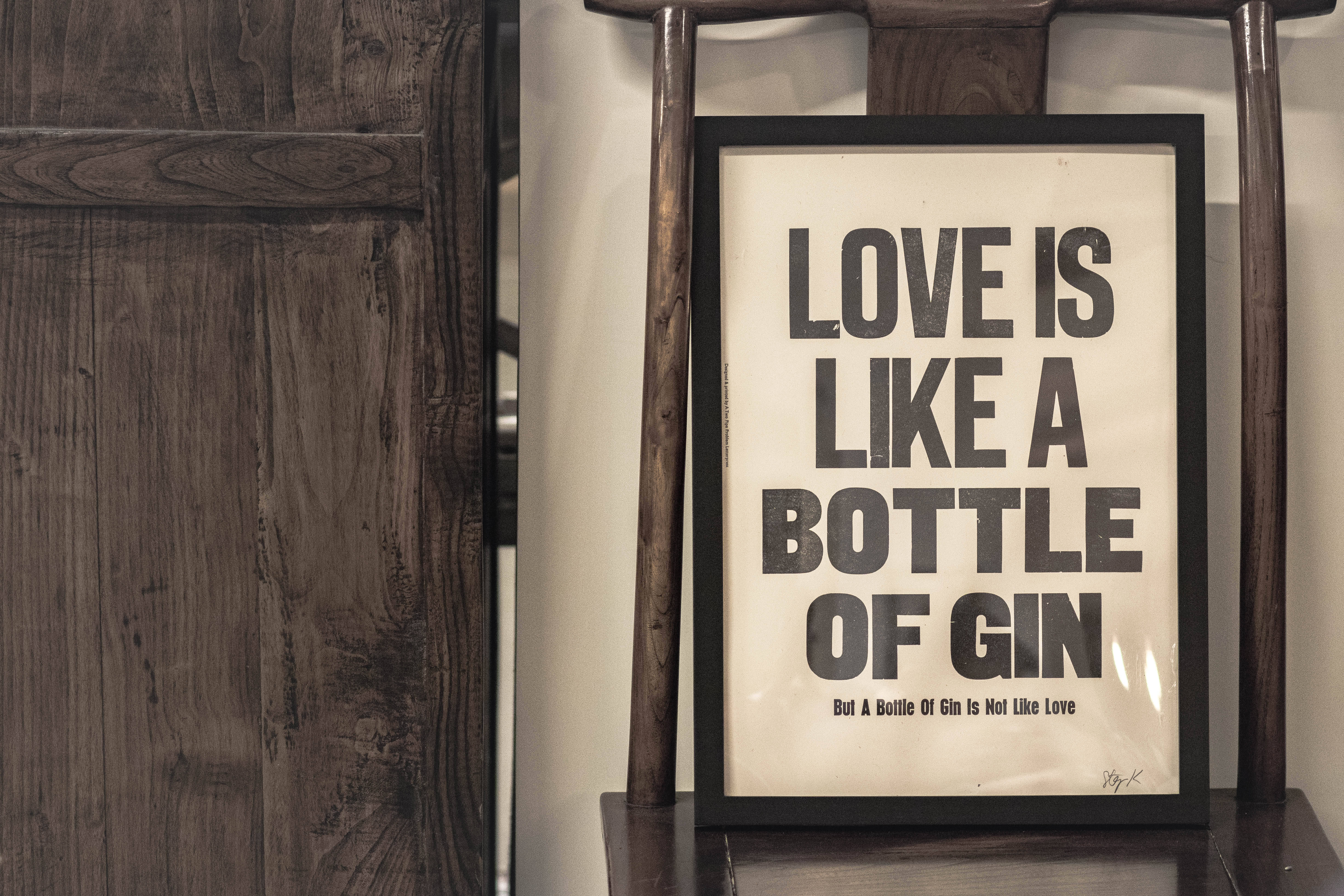 字型 桌布 壁紙 — 一個帶框的字體設計海報，上面寫著「LOVE IS LIKE A BOTTLE OF GIN」，放在一張木椅上，旁邊是質樸的木鑲板牆。