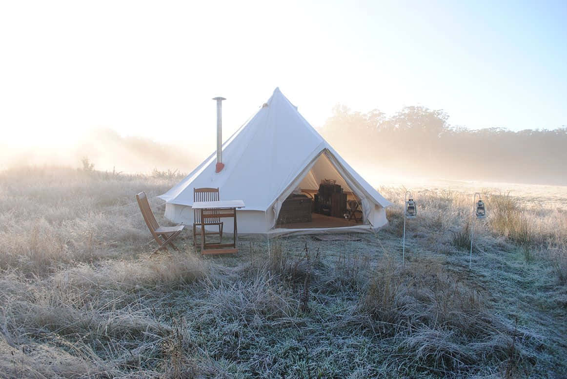 Winter Camping 桌布 壁紙 — 一個帶煙囪的白色鐘形帳篷矗立在霜凍的田野中，日出時分，周圍霧氣升騰。