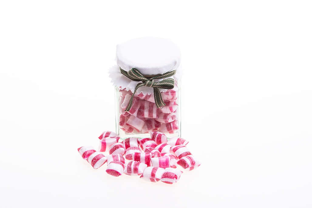 糖果 桌布 壁紙 — 一個帶白色蓋子和綠色蝴蝶結的玻璃罐，在純白色背景上裝滿了粉紅色和白色條紋的薄荷糖。