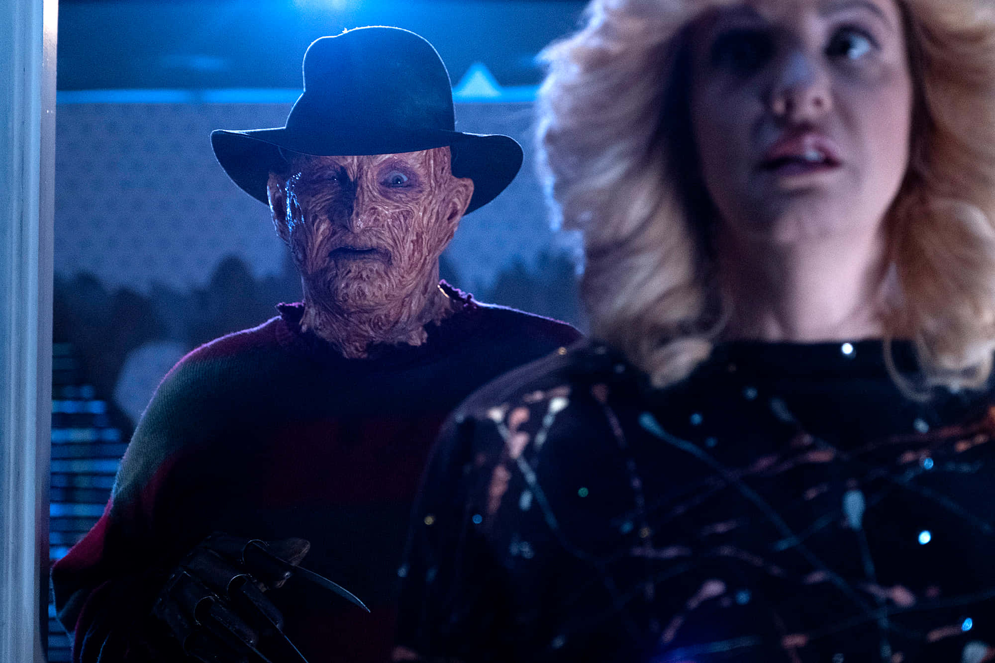 Freddy Krueger 桌布 壁紙 — 一個帶著標誌性帽子和毛衣的可怕佛萊迪·克魯格站在黑暗房間裡一個驚恐的金髮女人身後。