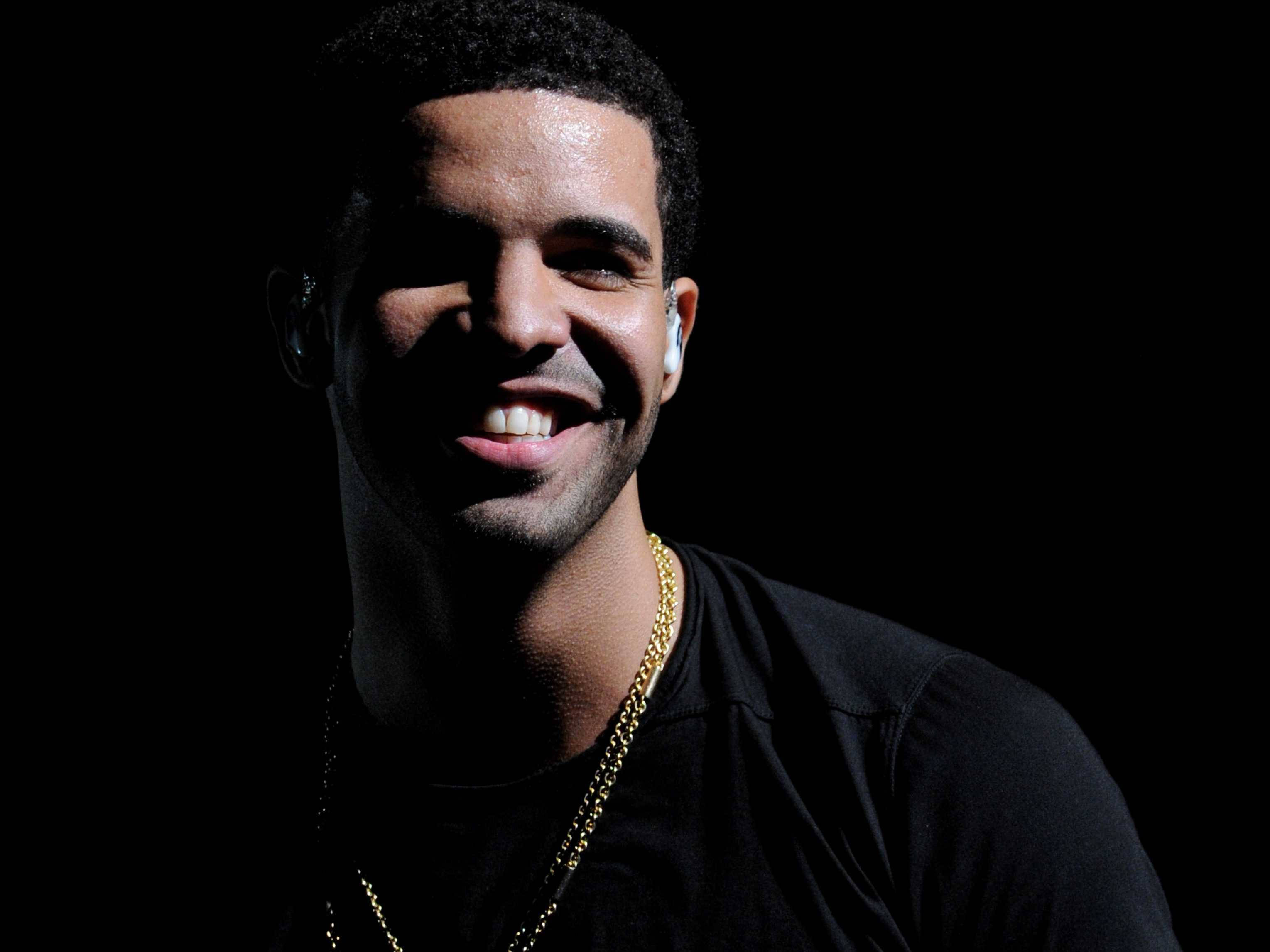 Drake 桌布 壁紙 — 一個帶著溫暖笑容和金色鏈條的男人，在戲劇性燈光下，在純黑背景中閃耀。