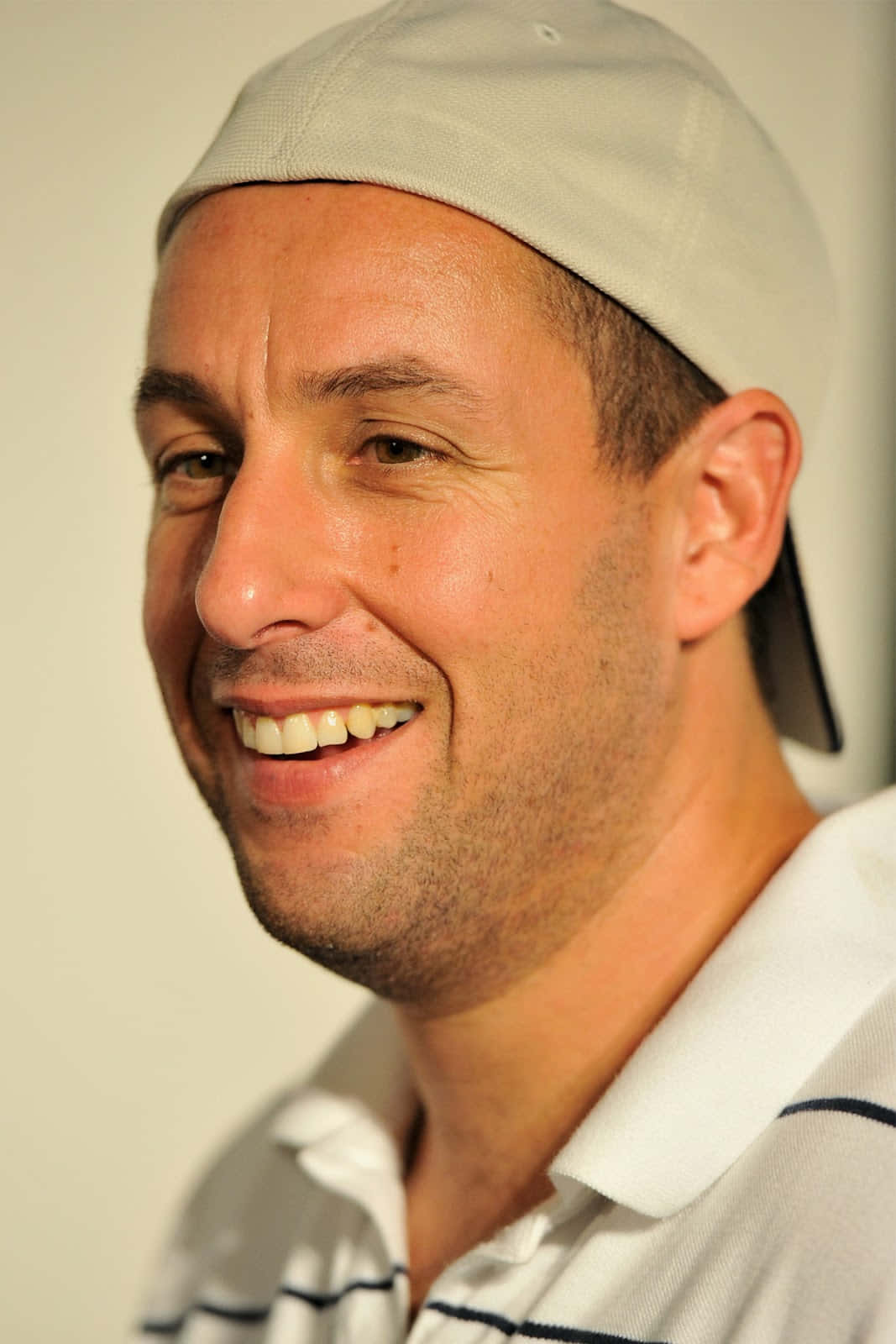 Adam Sandler 桌布 壁紙 — 一個帶著溫暖笑容的男人，在純色背景前戴著淺灰色帽子，穿著白色Polo衫。