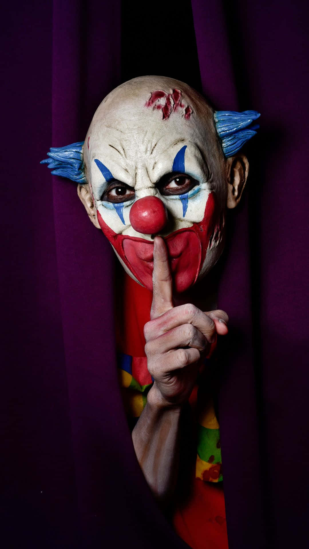 Horror Costumes 桌布 壁紙 — 一個帶血妝和藍色頭髮的威脅性小丑從紫色窗簾後窺視，手指放在嘴唇上做出無聲的威脅。