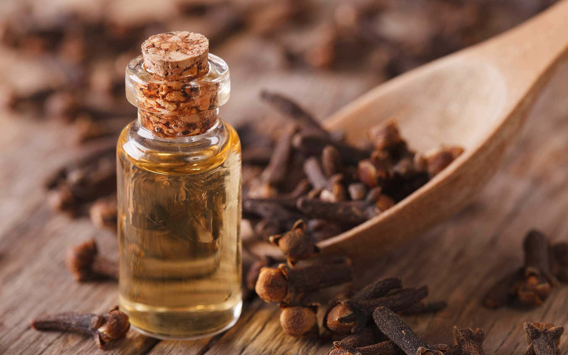 Essential Oil 桌布 壁紙 — 一個帶軟木塞的玻璃丁香精油瓶，旁邊放著一把盛滿整粒丁香的木勺，置於質樸的木質表面上。