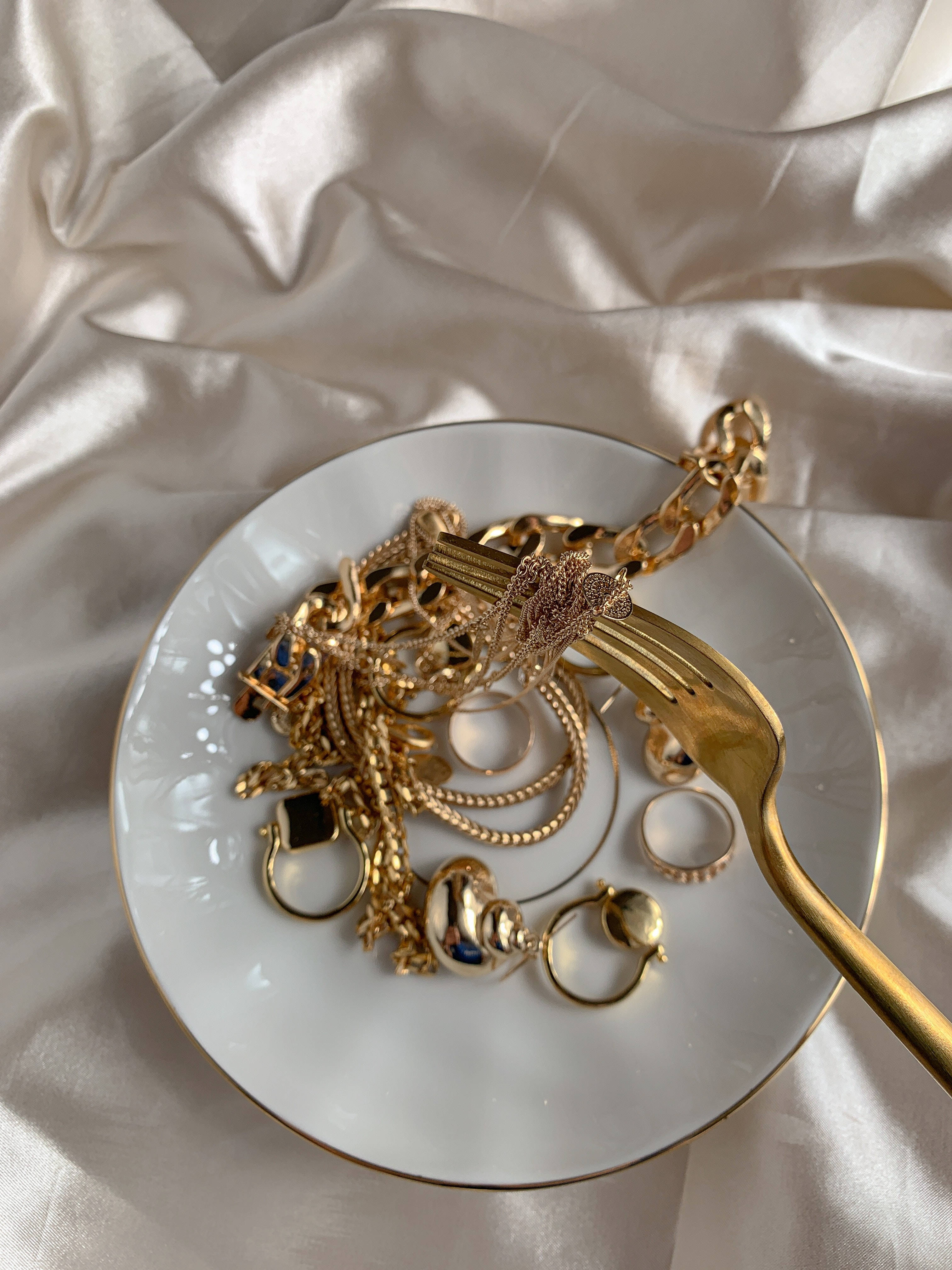 Gold Jewellery 桌布 壁紙 — 一個帶金邊的白色盤子，上面放著纏繞的金飾和叉子，置於皺巴巴的奶油色緞布上。