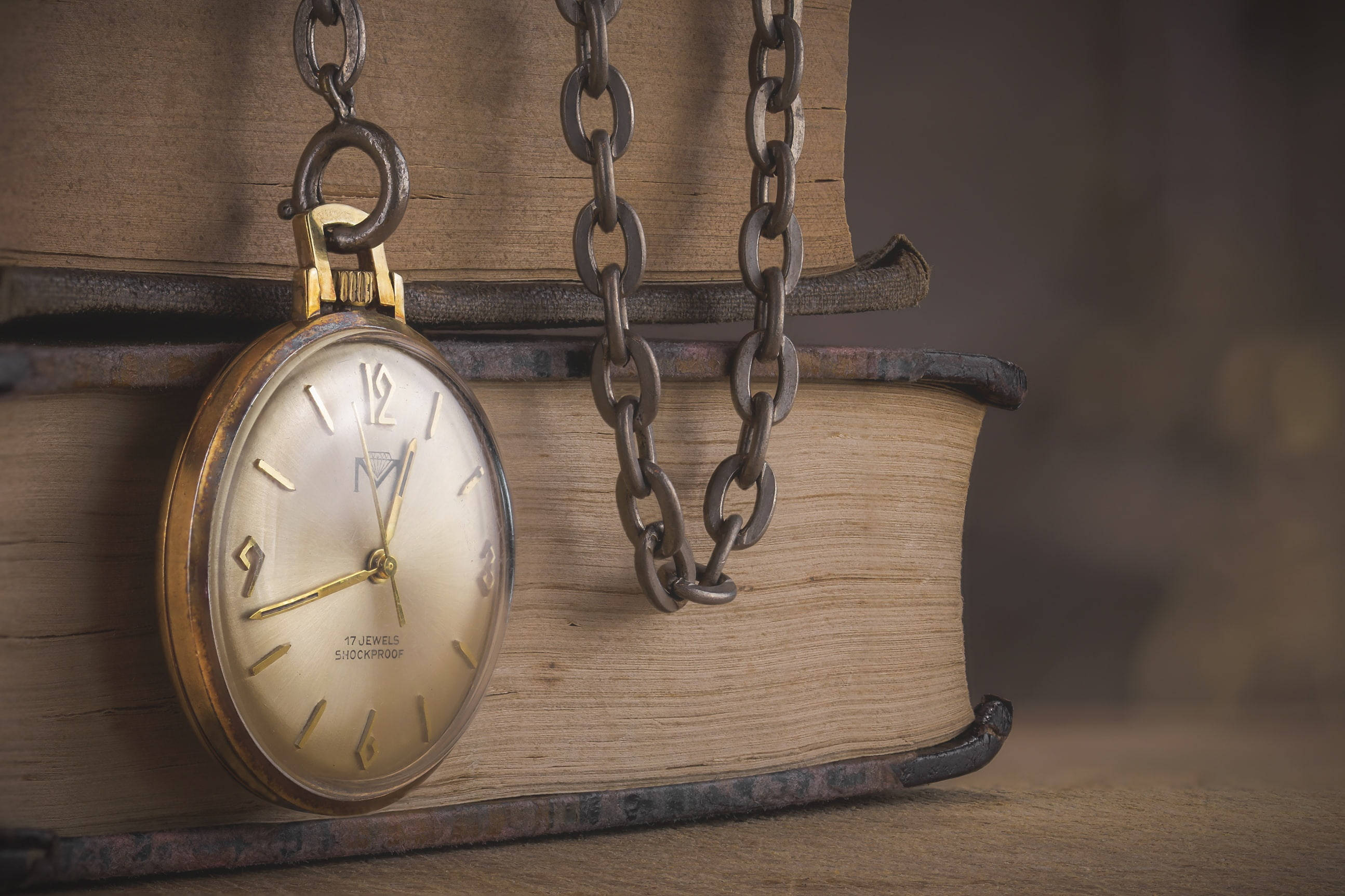 Clock 桌布 壁紙 — 一個帶鏈的復古金色懷錶擱在一本皮革裝訂磨損的舊書書脊上。