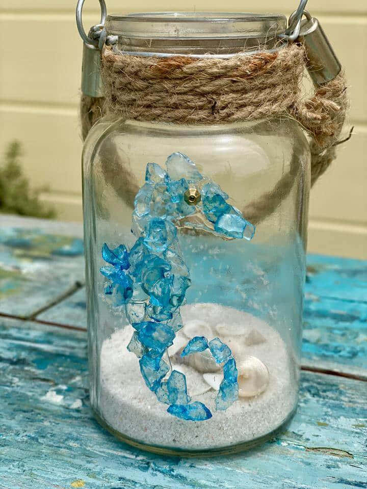 Sea Glass 桌布 壁紙 — 一個帶麻繩蓋的玻璃罐，裡面裝著藍色水晶海馬和貝殼在沙中，放在風化的藍色木桌上。