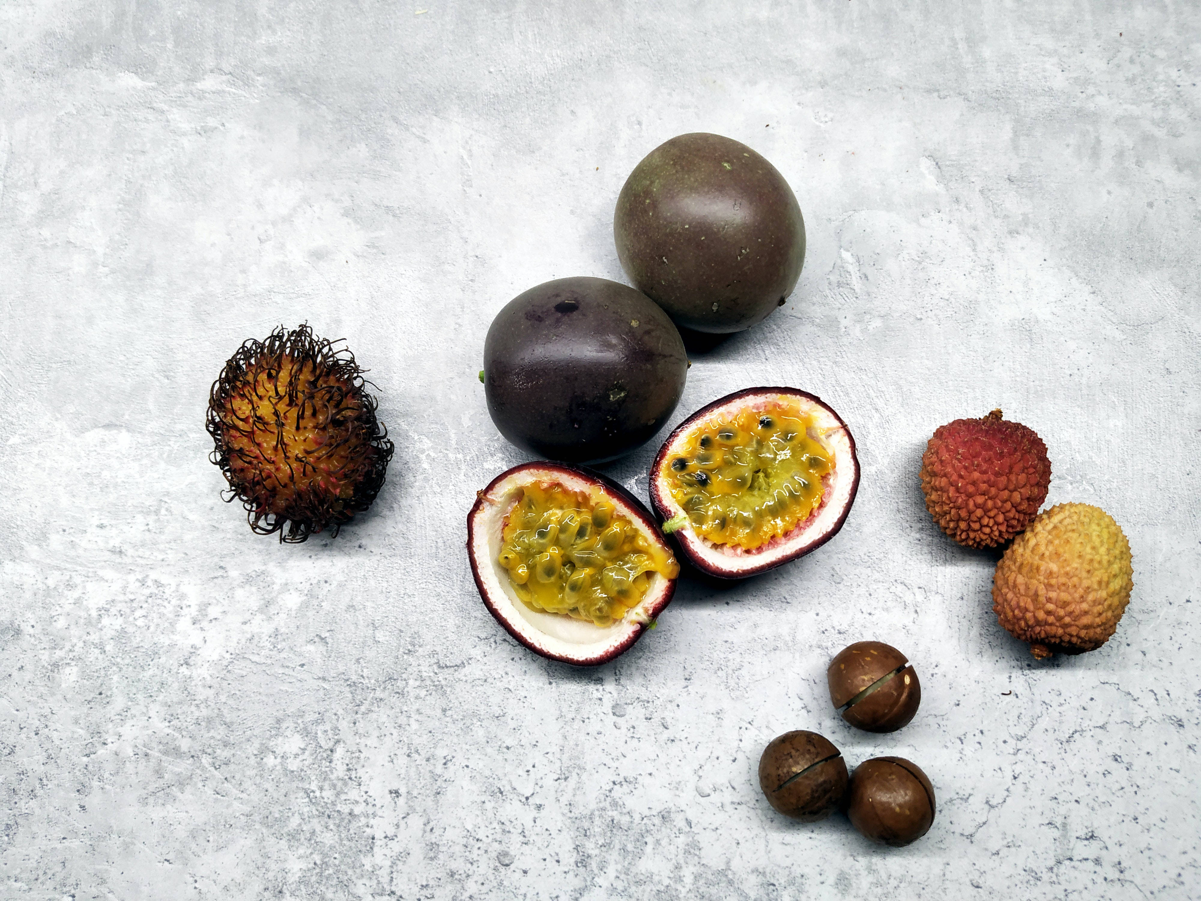 Litchi 桌布 壁紙 — 一個平鋪的異國熱帶水果擺設，包括紅毛丹、荔枝、百香果和夏威夷果仁，放在有紋理的灰色表面上。