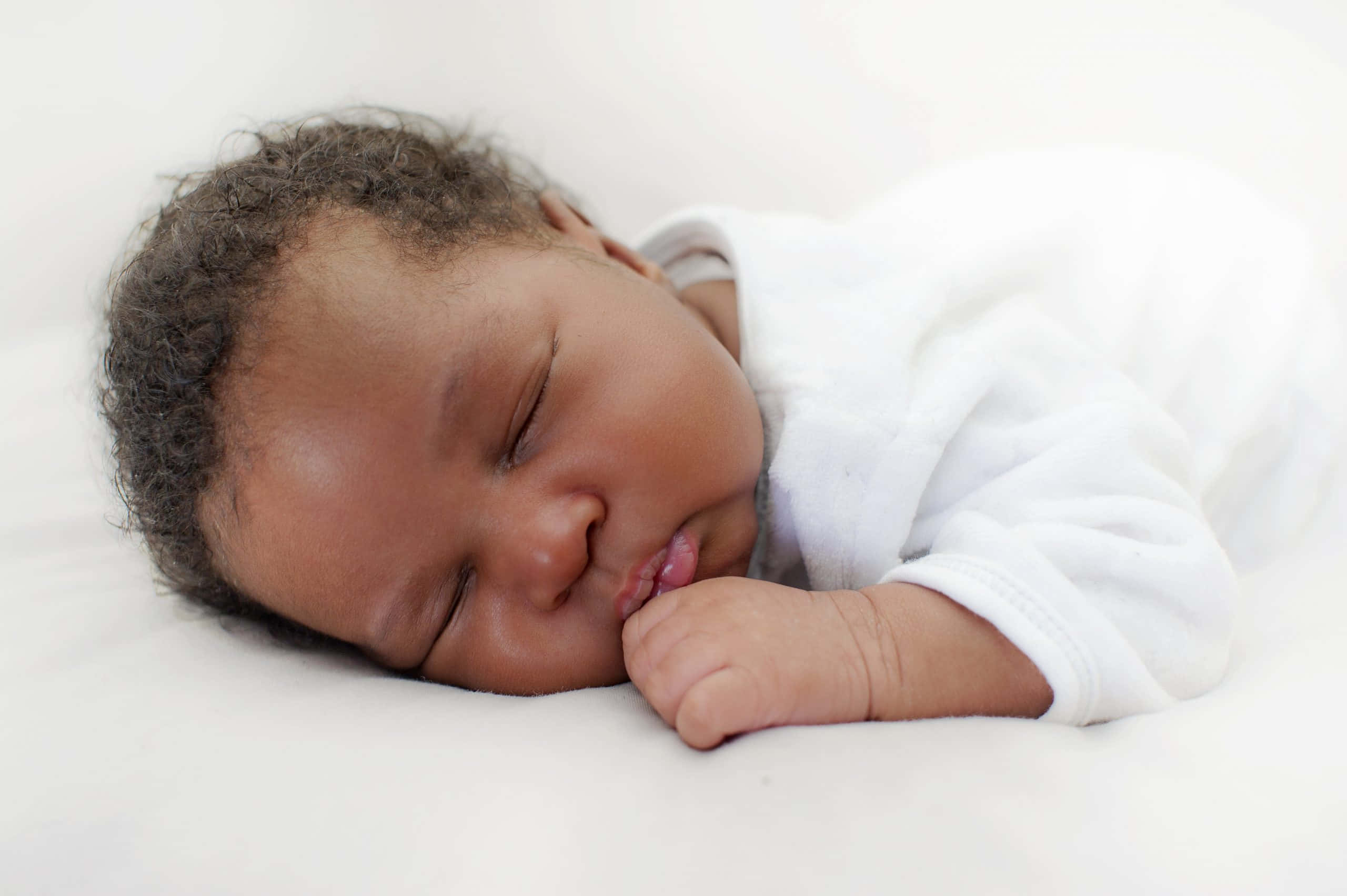 Newborn Baby 桌布 壁紙 — 一個平靜的新生嬰兒，深色捲髮，在柔軟的白色毯子上安睡，小拳頭靠近嘴唇。