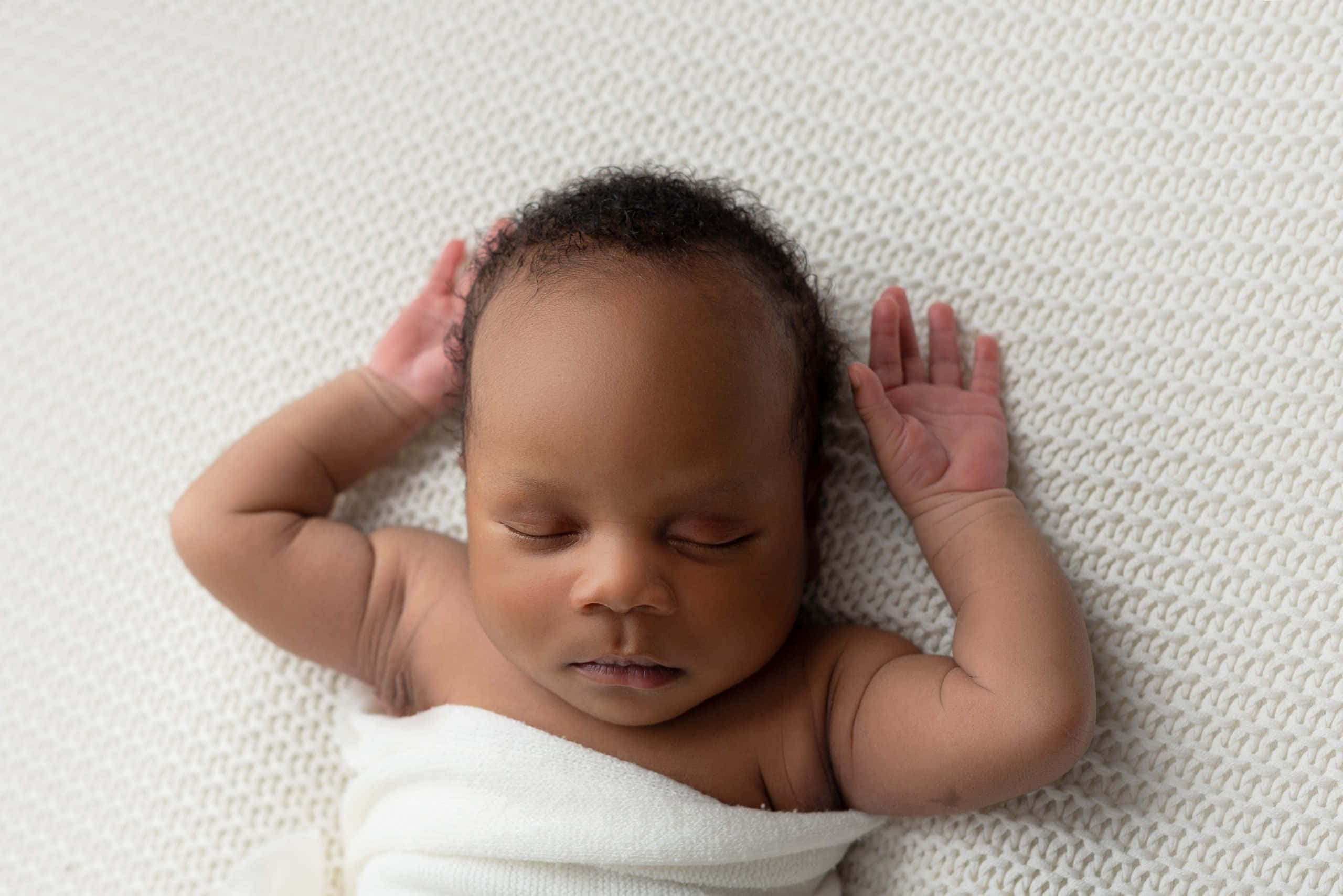Newborn Baby 桌布 壁紙 — 一個平靜的新生嬰兒，深色皮膚和捲曲頭髮，被白色包裹著，在帶有紋理的奶油色背景上安睡。