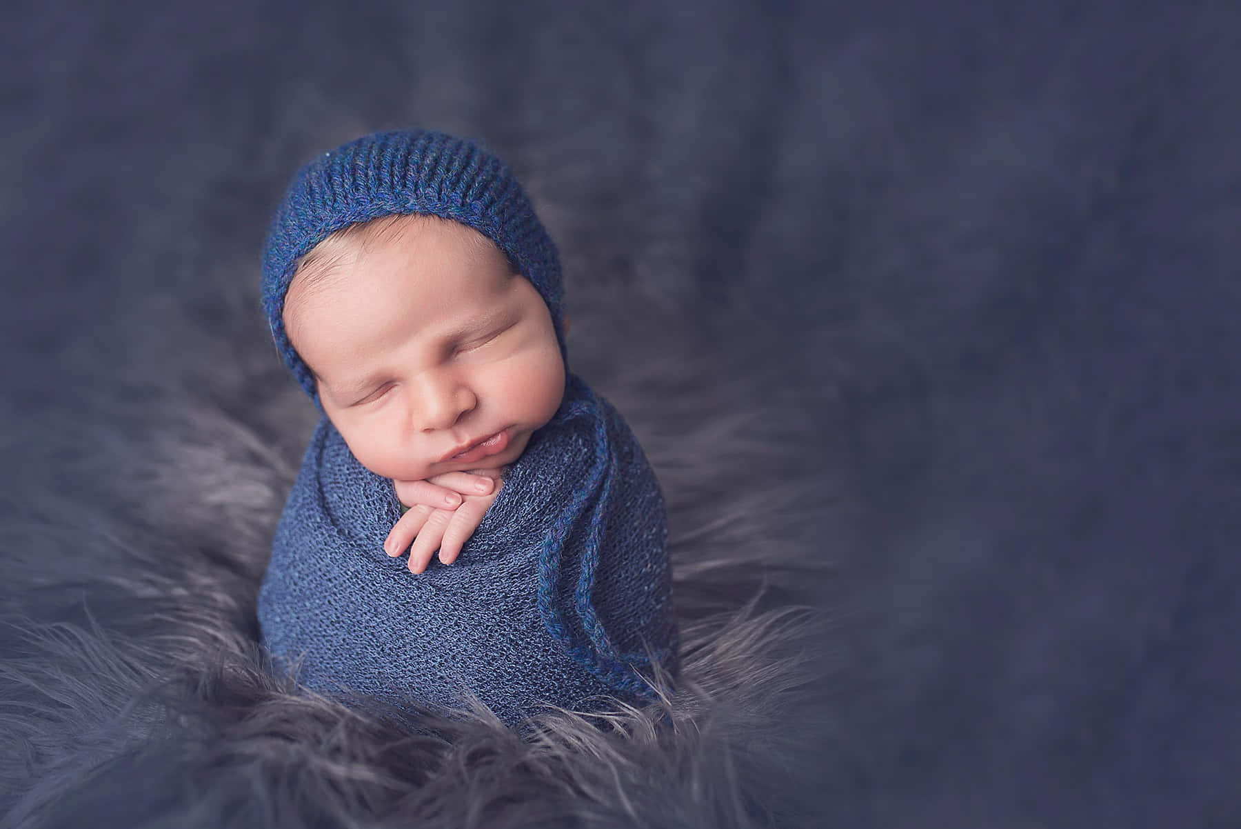 Newborn Baby 桌布 壁紙 — 一個平靜的新生嬰兒包裹在藍色針織布料中，安詳地睡在柔軟的灰色毛毯上。