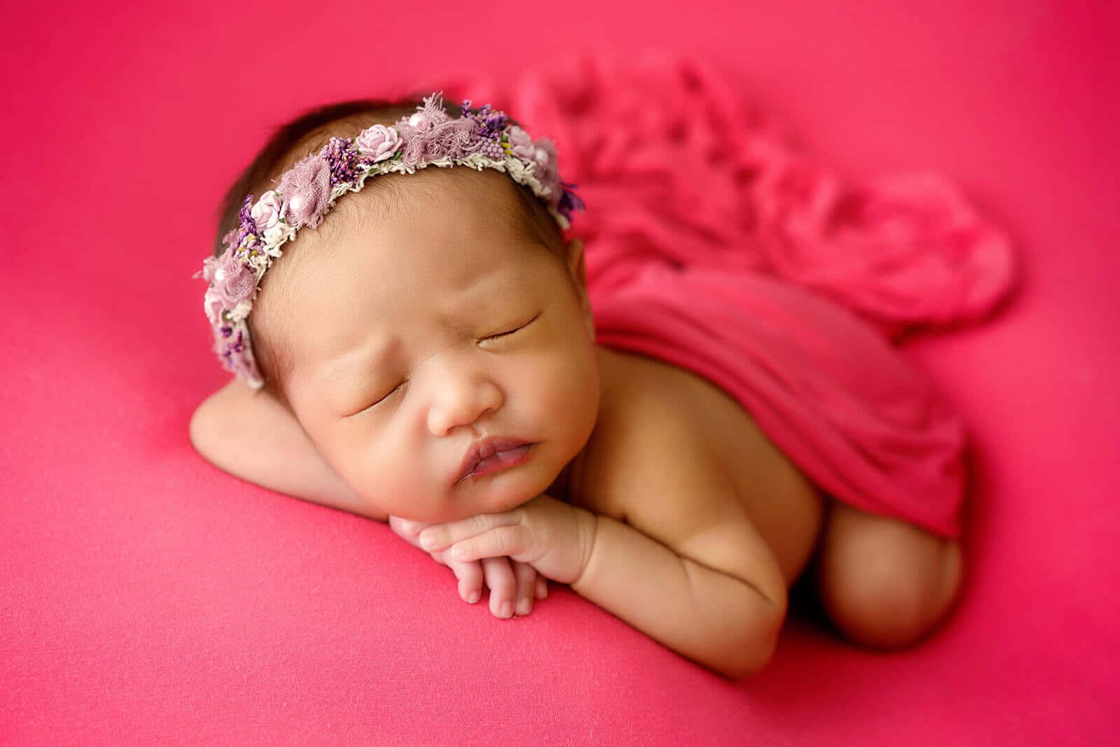 Newborn Baby 桌布 壁紙 — 一個平靜的新生嬰兒在充滿活力的粉紅色布料上安睡，並飾有精緻的花卉頭帶。