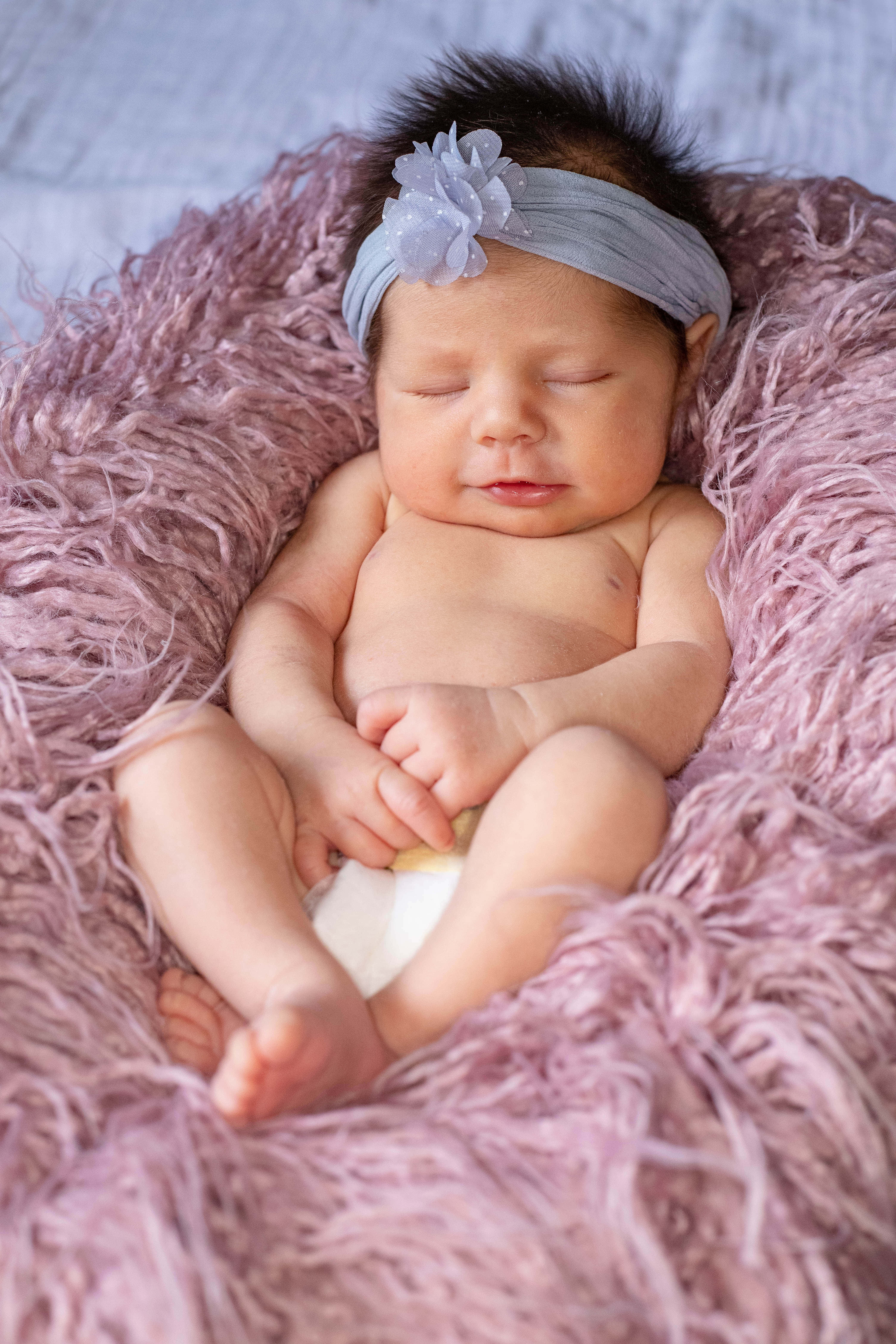 Newborn Baby 桌布 壁紙 — 一個平靜的新生嬰兒在柔軟蓬鬆的薰衣草色毛毯上安睡，戴著淺藍色花卉頭帶。