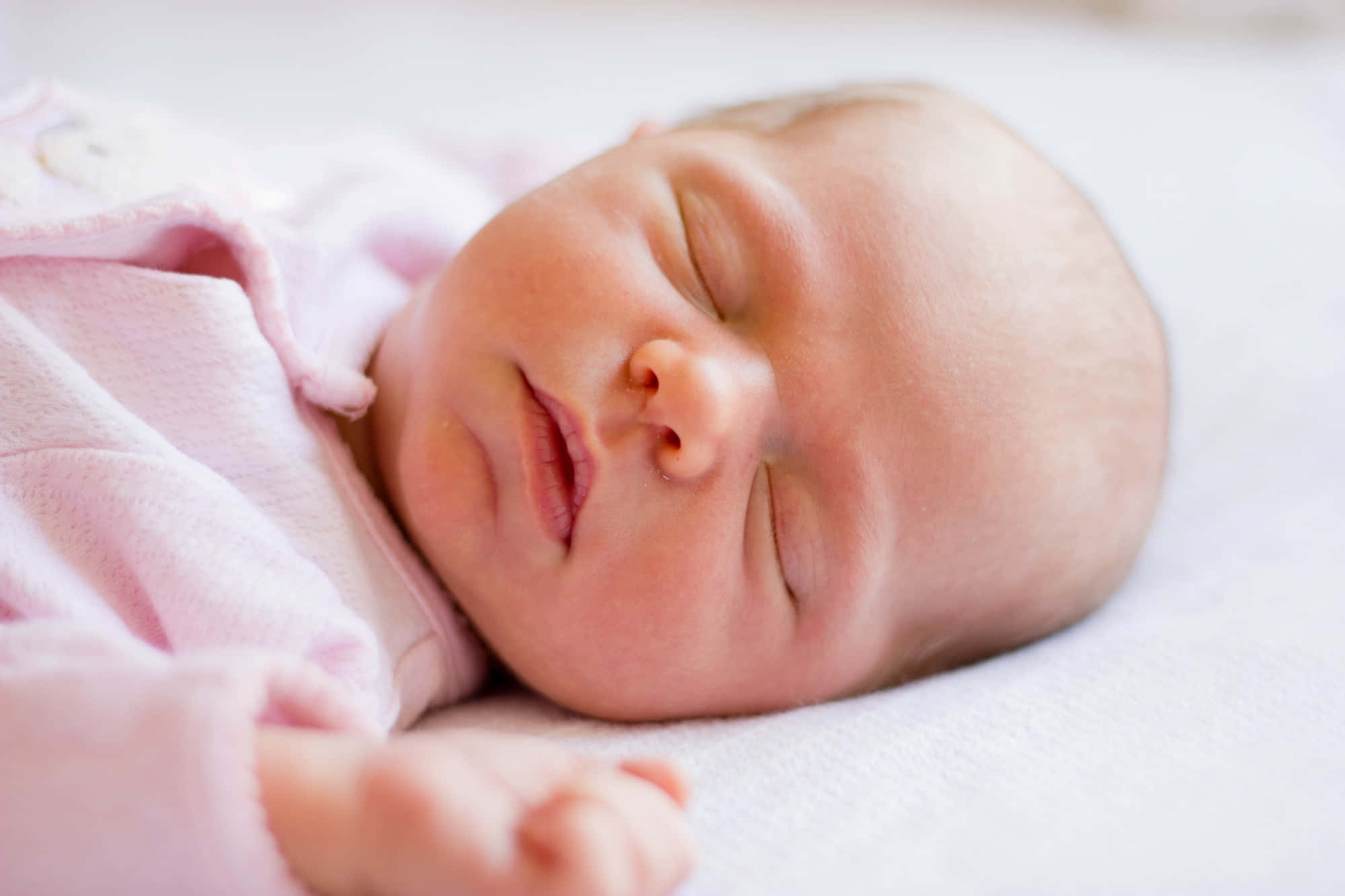 Infant 桌布 壁紙 — 一個平靜的新生嬰兒穿著柔軟的粉紅色連身衣，在白色毯子上安穩地睡著。