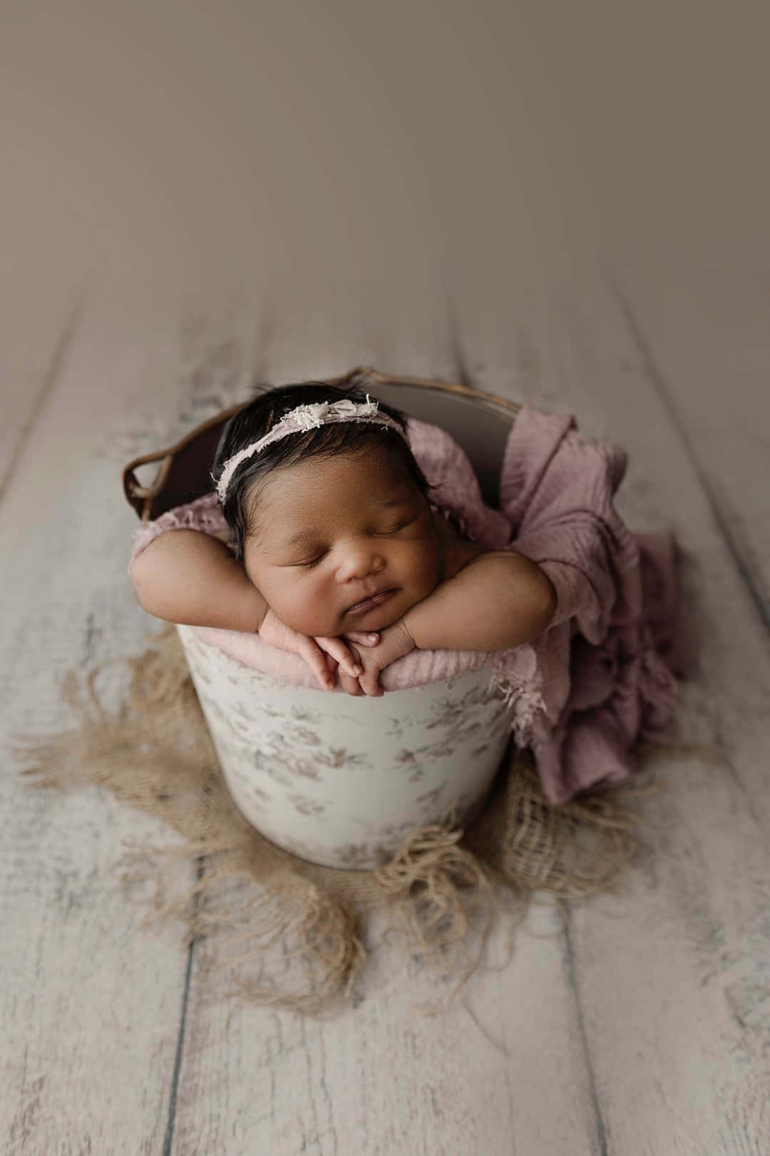 Newborn Baby 桌布 壁紙 — 一個平靜的新生嬰兒蜷縮在一個花卉桶中，包裹在柔軟的淡紫色布料裡，躺在白色粉刷的木地板上睡覺。