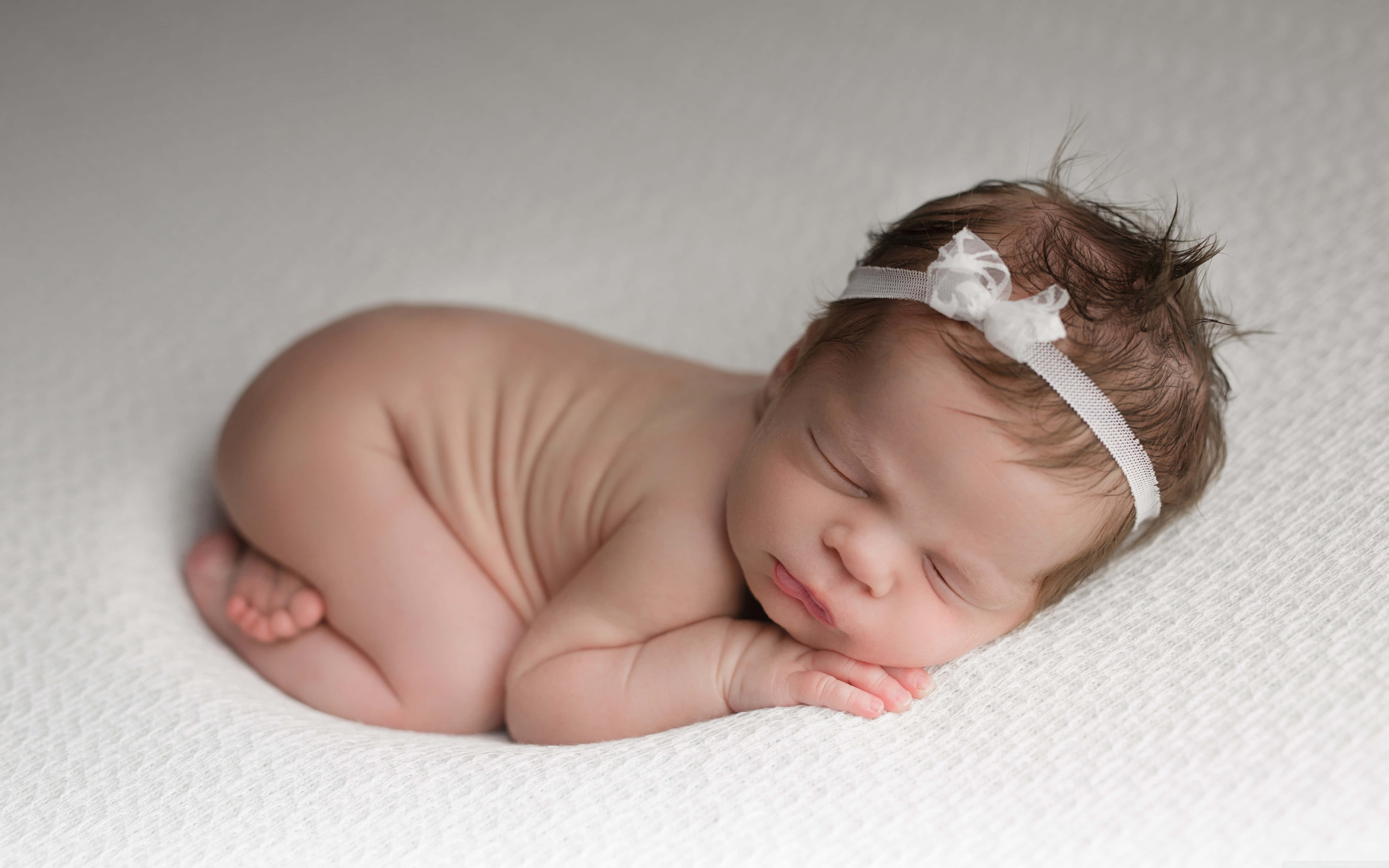 Baby Girl 桌布 壁紙 — 一個平靜的新生嬰兒蜷縮在柔軟的白色紋理毛毯上睡覺，戴著精緻的白色頭帶。